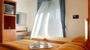 Habitación de hotel con cama, televisor en un estante, mesita de noche con un jarrón y ventana con cortinas.