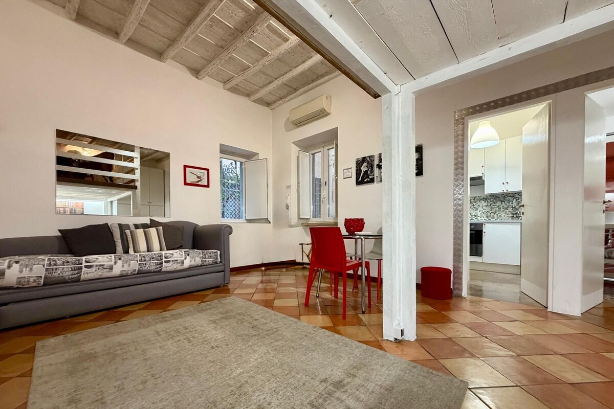 A living room with a sofa, a red chair, a rug, and a kitchen visible through an open door.