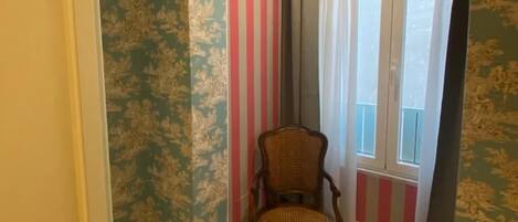 A room with patterned wallpaper, a wooden chair, a window with striped curtains, and a ceiling light.