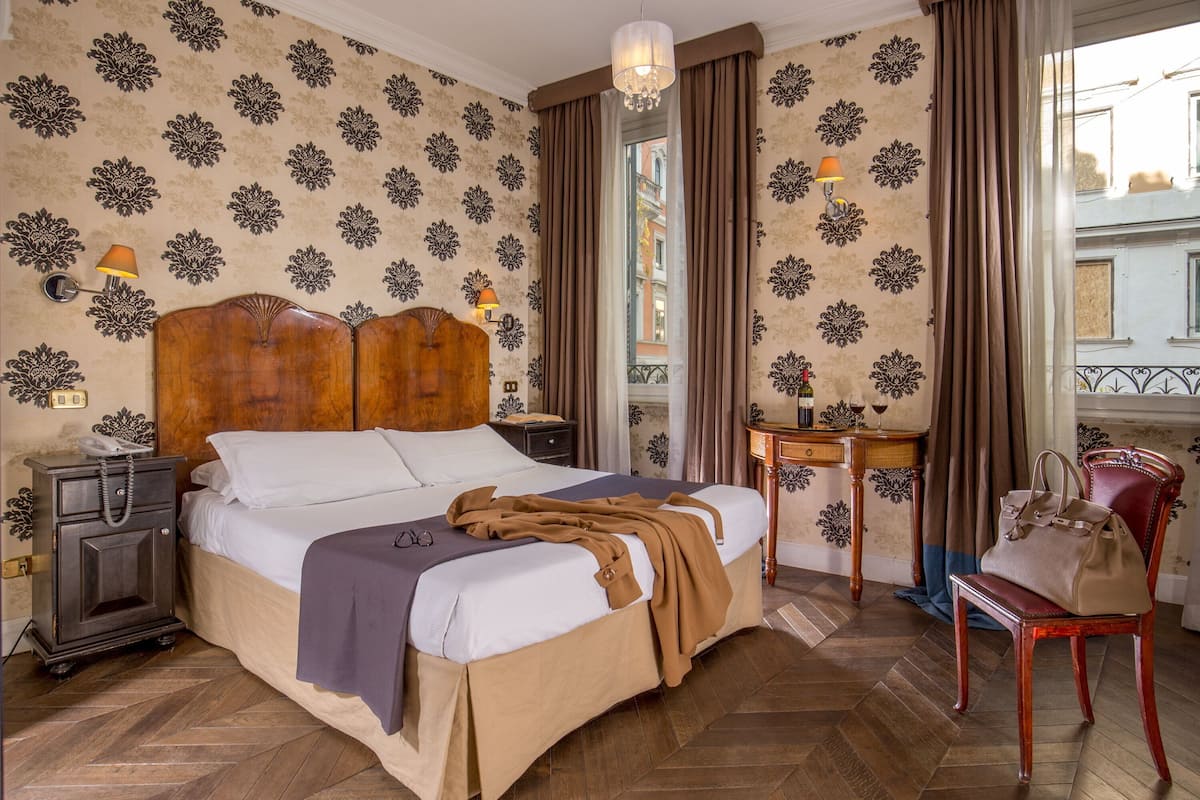 A hotel room with a large bed, wooden headboard, patterned wallpaper, a wooden nightstand, a bedside table with a lamp, and a view of the city through the window.