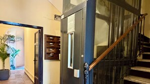 Uma escadaria com corrimão preto e porta de vidro, que dá acesso a um quarto com piso ladrilhado e planta em vaso.