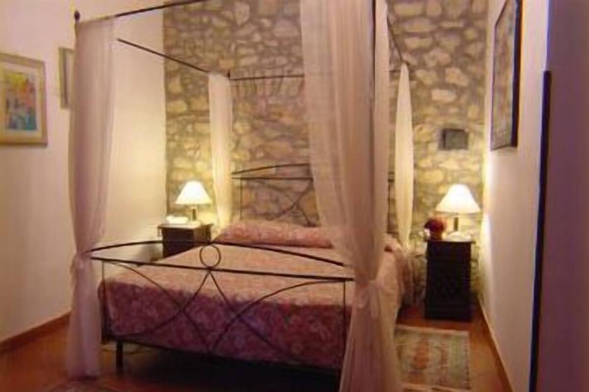A four-poster bed with a stone wall background, two bedside tables with lamps, and a patterned rug on the floor.