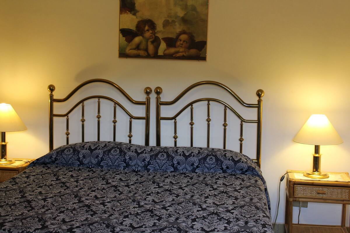 A bed with a patterned bedspread, two bedside lamps, and a painting of cherubs on the wall.