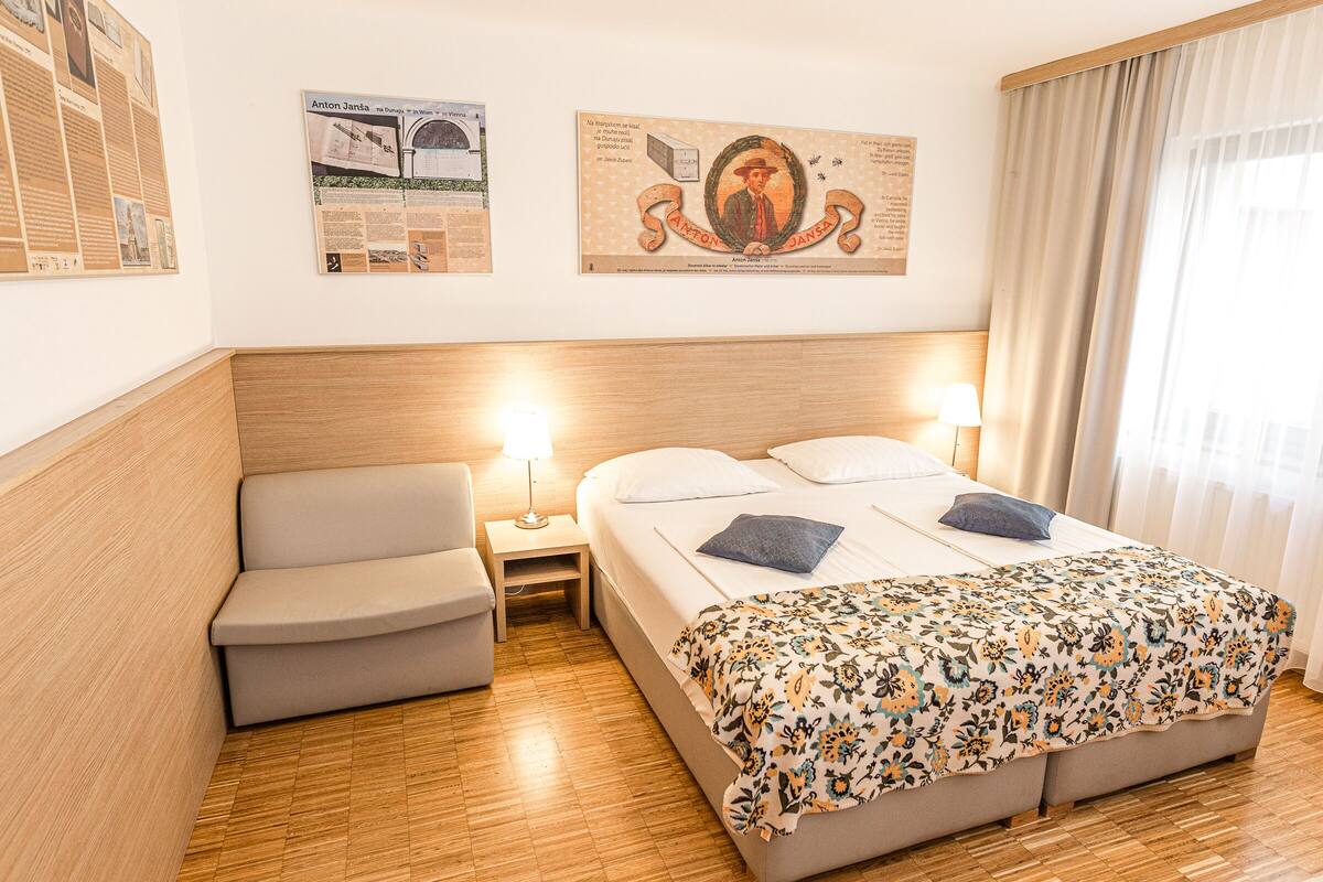 Una camera d'albergo con un letto, una sedia, un comodino e due poster informativi appesi al muro.