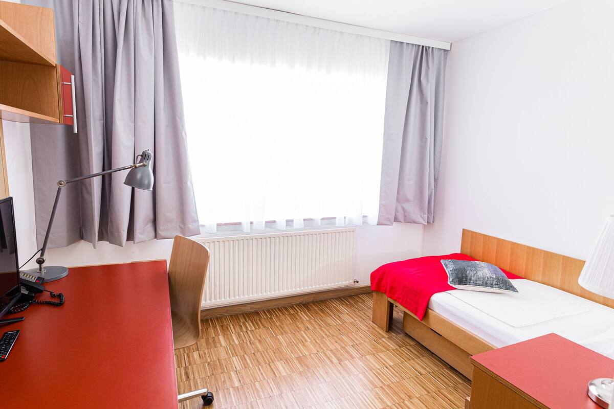 Una camera d'albergo con un letto, una scrivania, una sedia, una lampada e una finestra con tende.