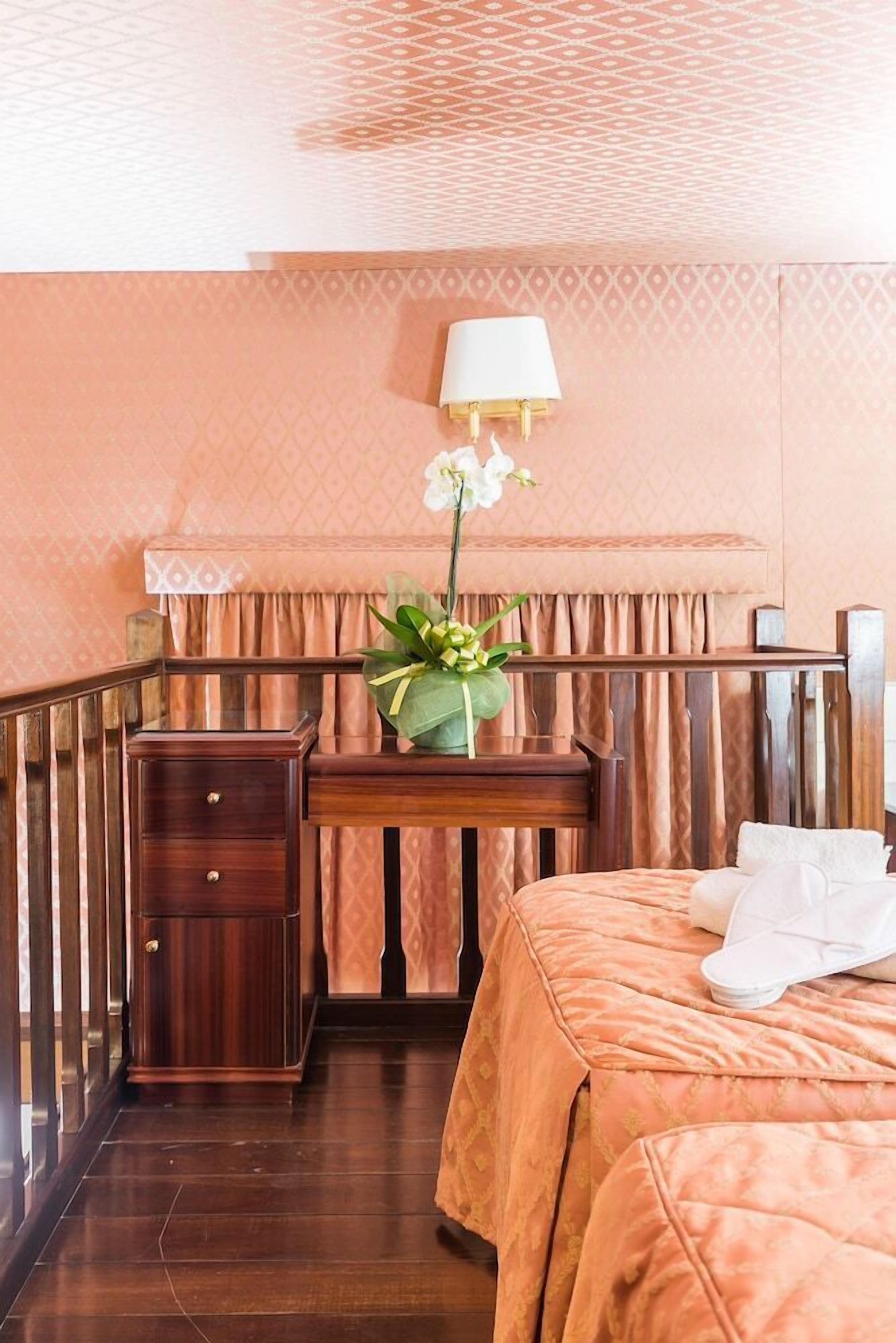Une chambre d’hôtel avec un lit, une table de chevet en bois, un bureau, une lampe et un vase avec des fleurs.