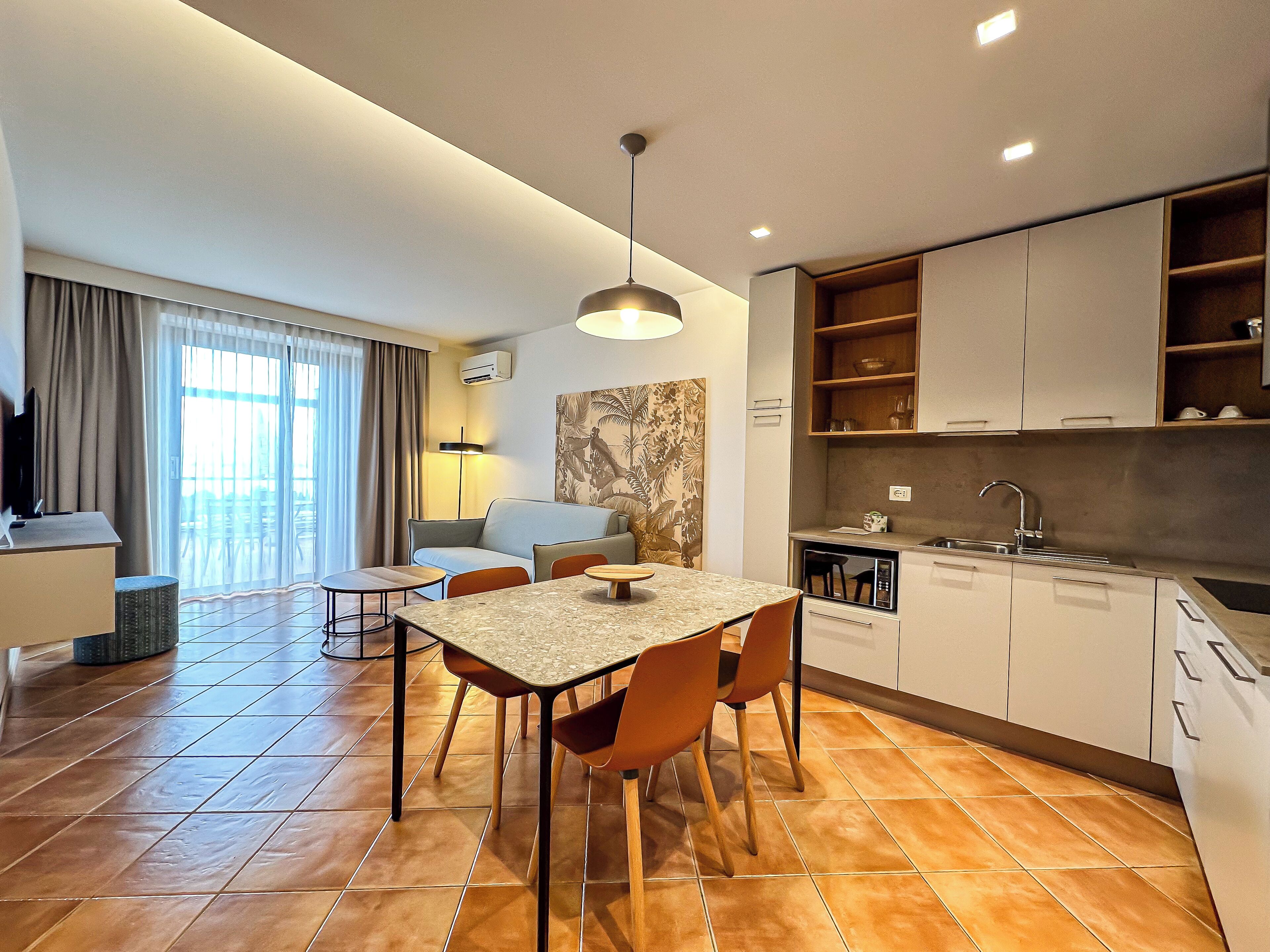 A modern hotel room with a dining area, kitchenette, and a large window with curtains.