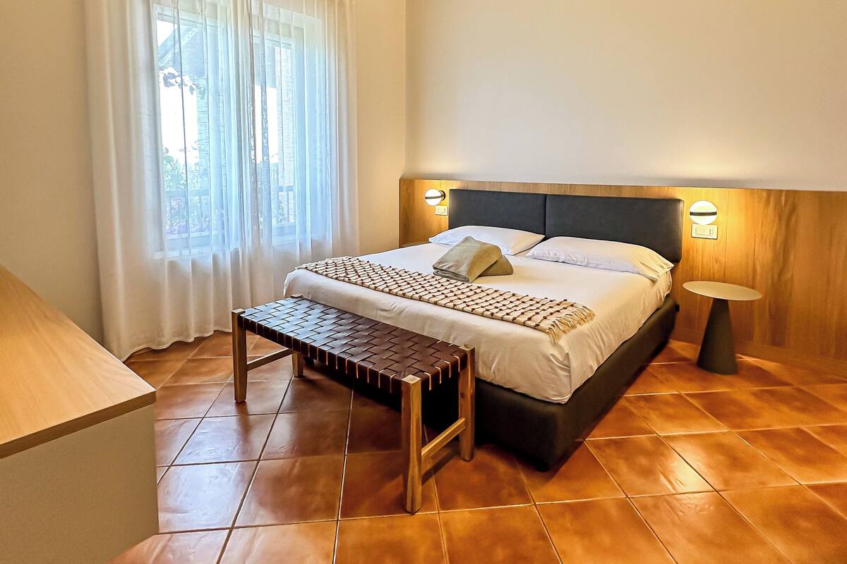 A bedroom with a bed, bench, bedside table, and a window with curtains.