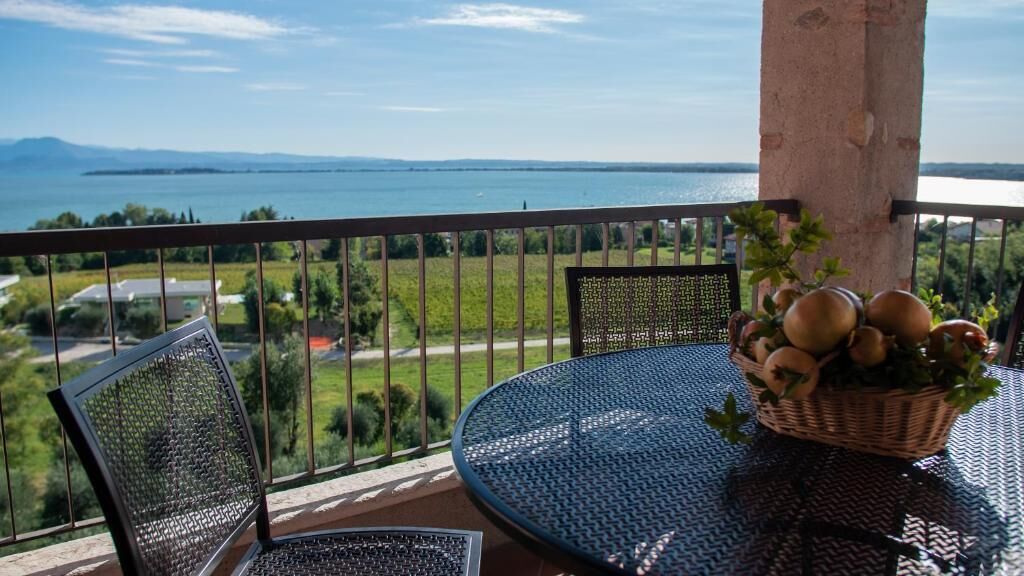 Un balcone con un tavolo rotondo e sedie, un cesto di vimini con della frutta, e vista su un'acqua e del verde.