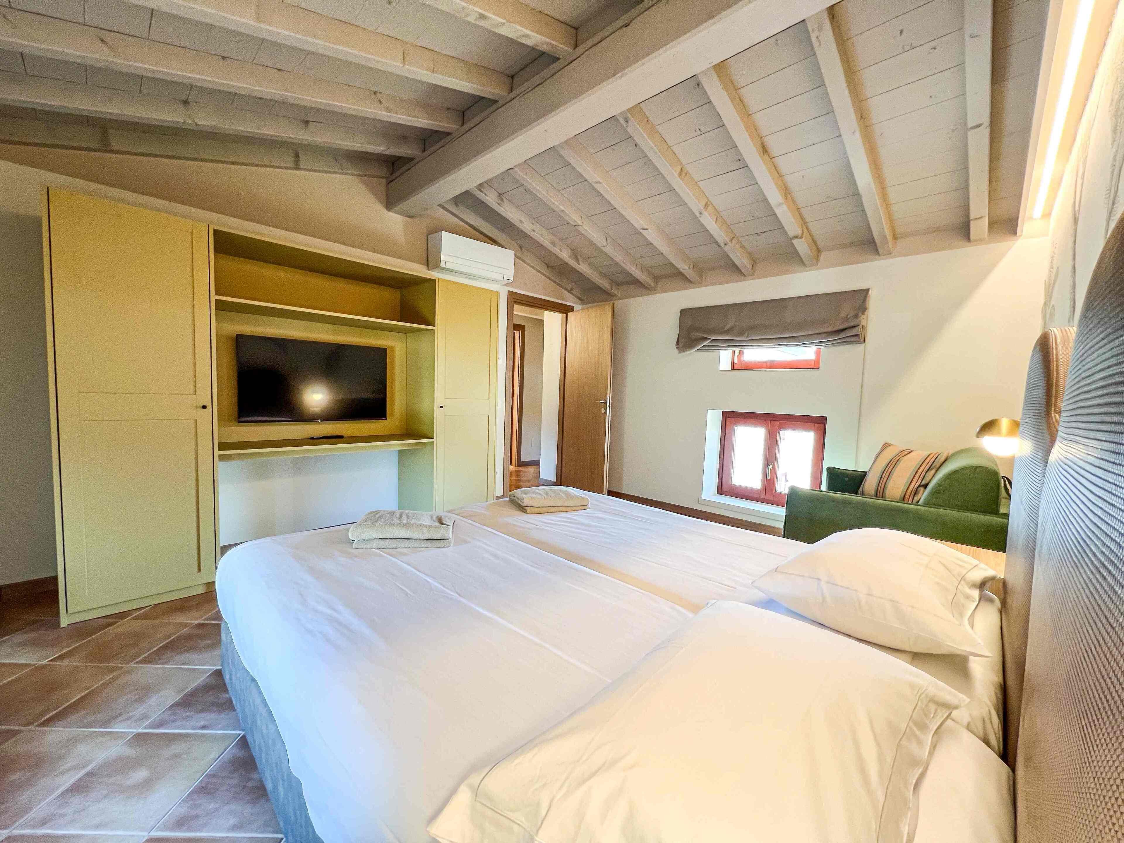 Een slaapkamer met een groot bed, een televisie op een plank, een raam en een groene fauteuil.