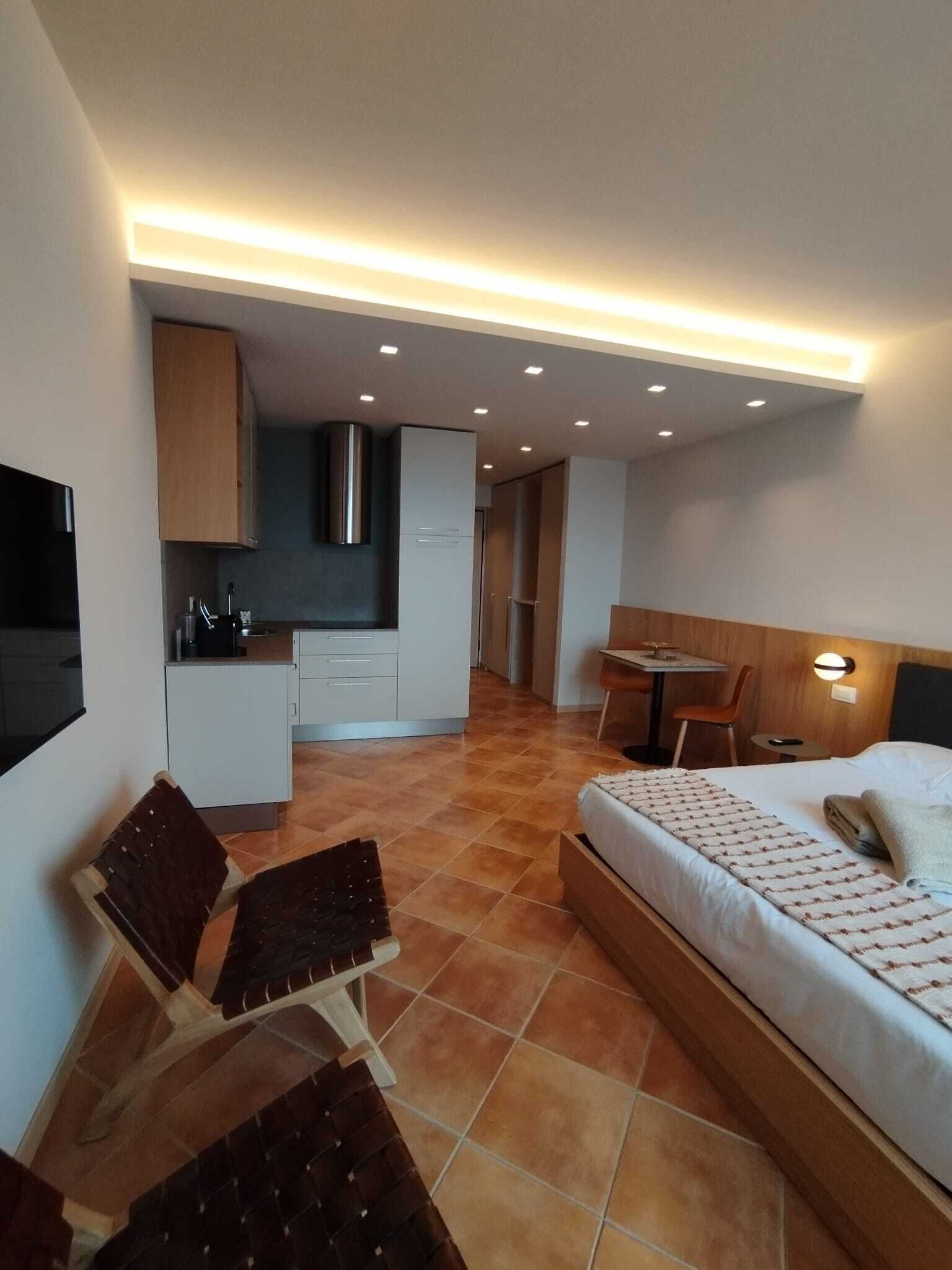 A compact hotel room with a bed, a small desk, a chair, a kitchenette, and a TV.