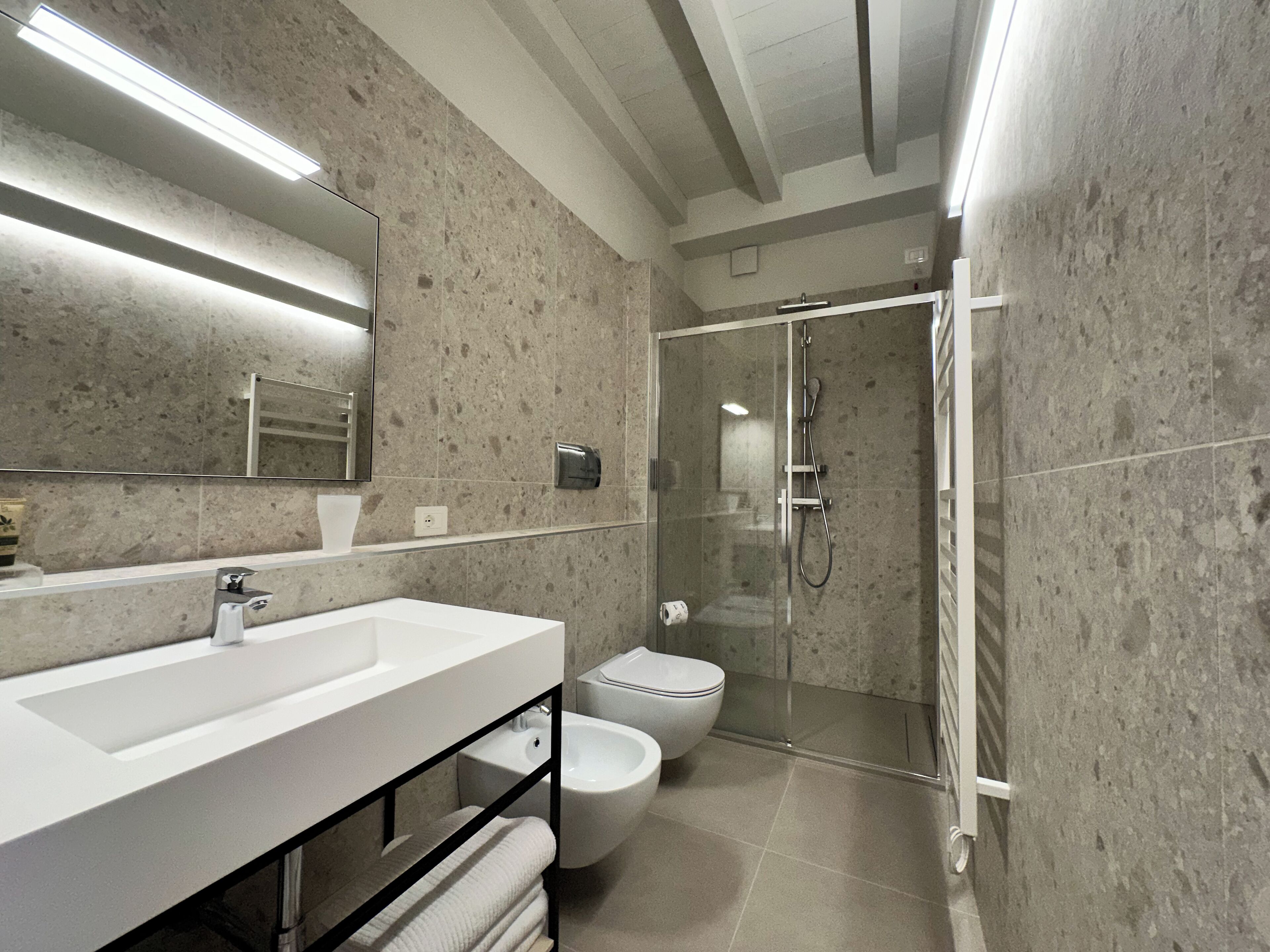 Une salle de bain moderne avec un grand miroir, un lavabo blanc, des toilettes et une douche séparée par une paroi de verre.