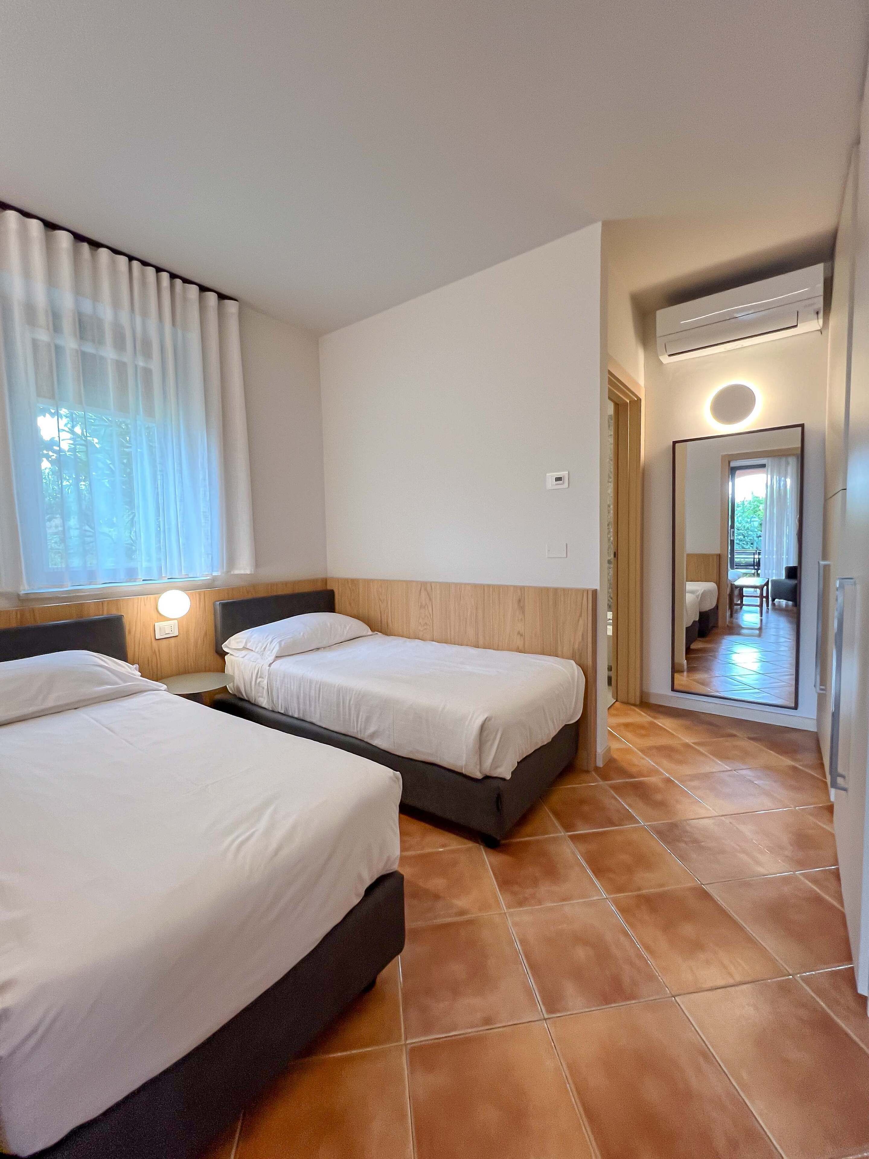 Habitación de hotel con dos camas, una mesita redonda, un espejo y vista al jardín por la ventana.
