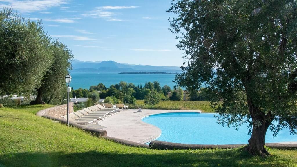 Une piscine à bord incurvé, entourée de pelouse et d’arbres, offrant une vue sur la mer et les montagnes lointaines.