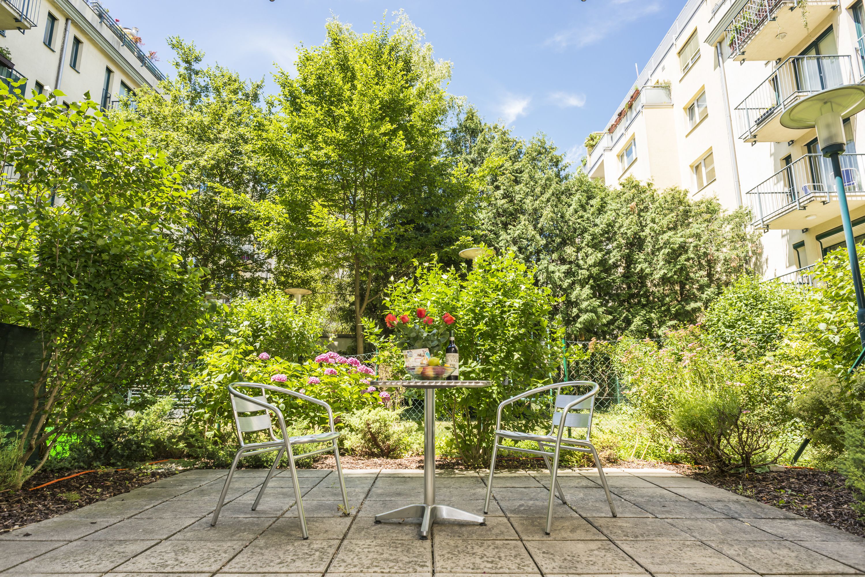 A garden patio with a table and chairs, surrounded by lush greenery and a building in the background.