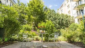 A garden patio with a table and chairs, surrounded by lush greenery and a building in the background.
