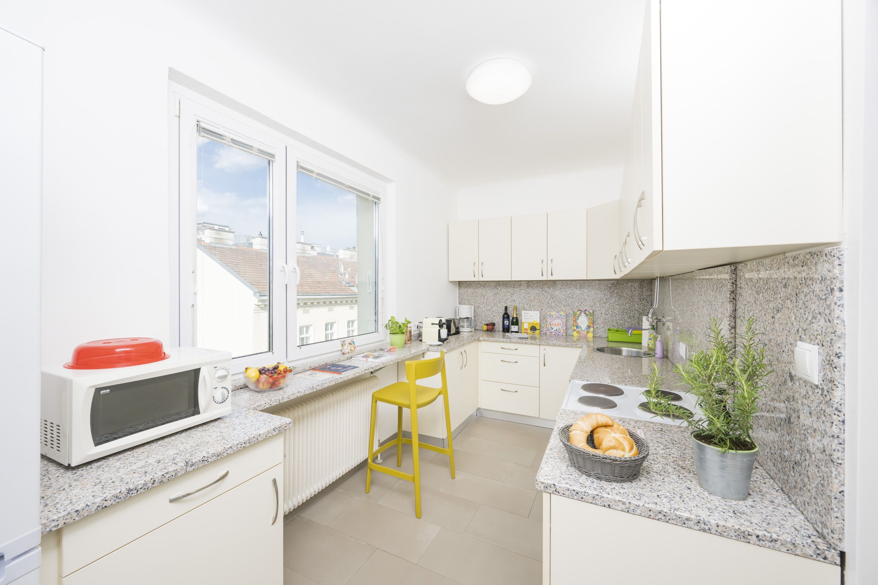 A modern kitchen with white cabinets, a red microwave, a yellow chair, a window with a view of buildings, and a potted plant.
