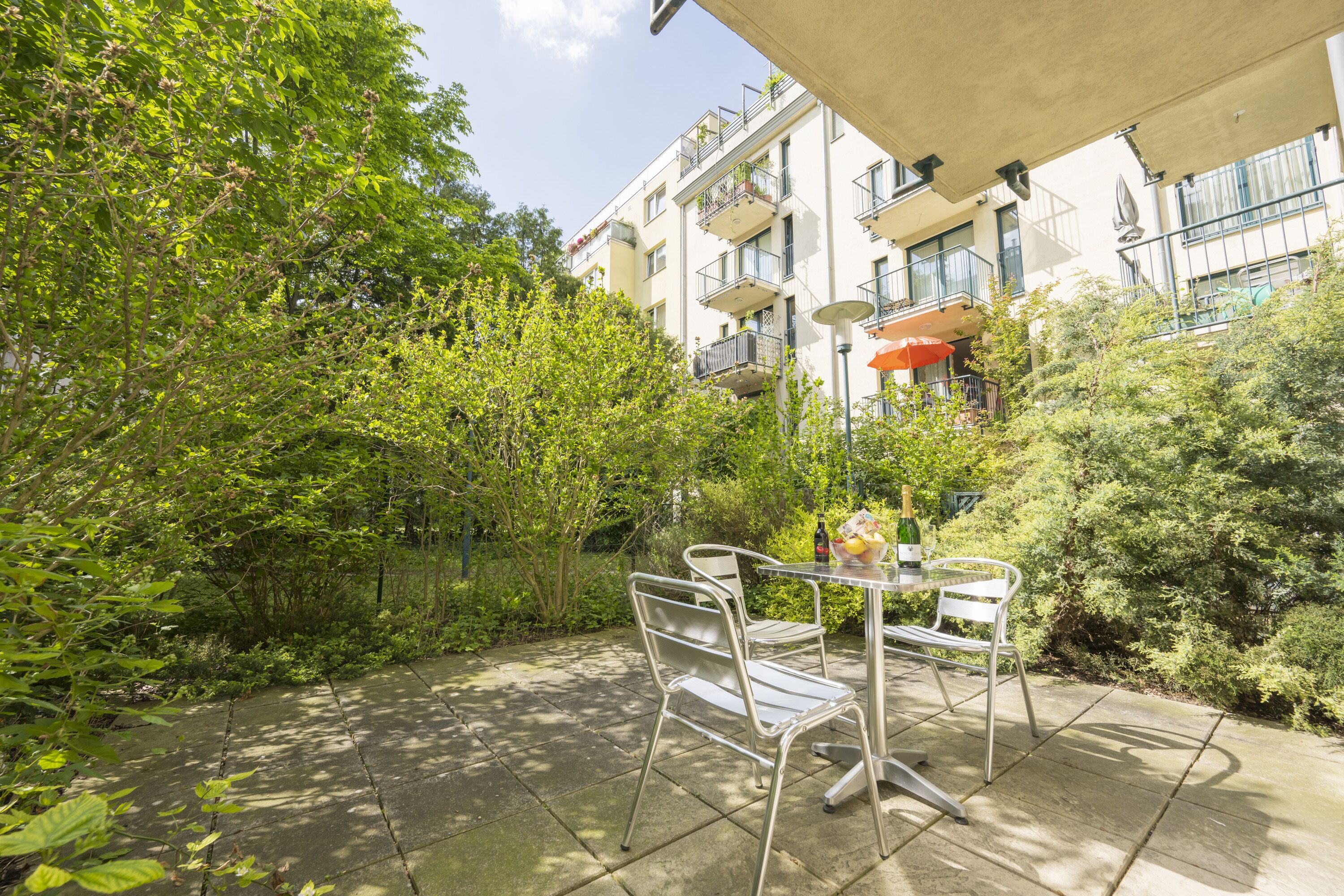 Un patio con sillas y una mesa de metal, rodeado de exuberante vegetación y un edificio con balcones.