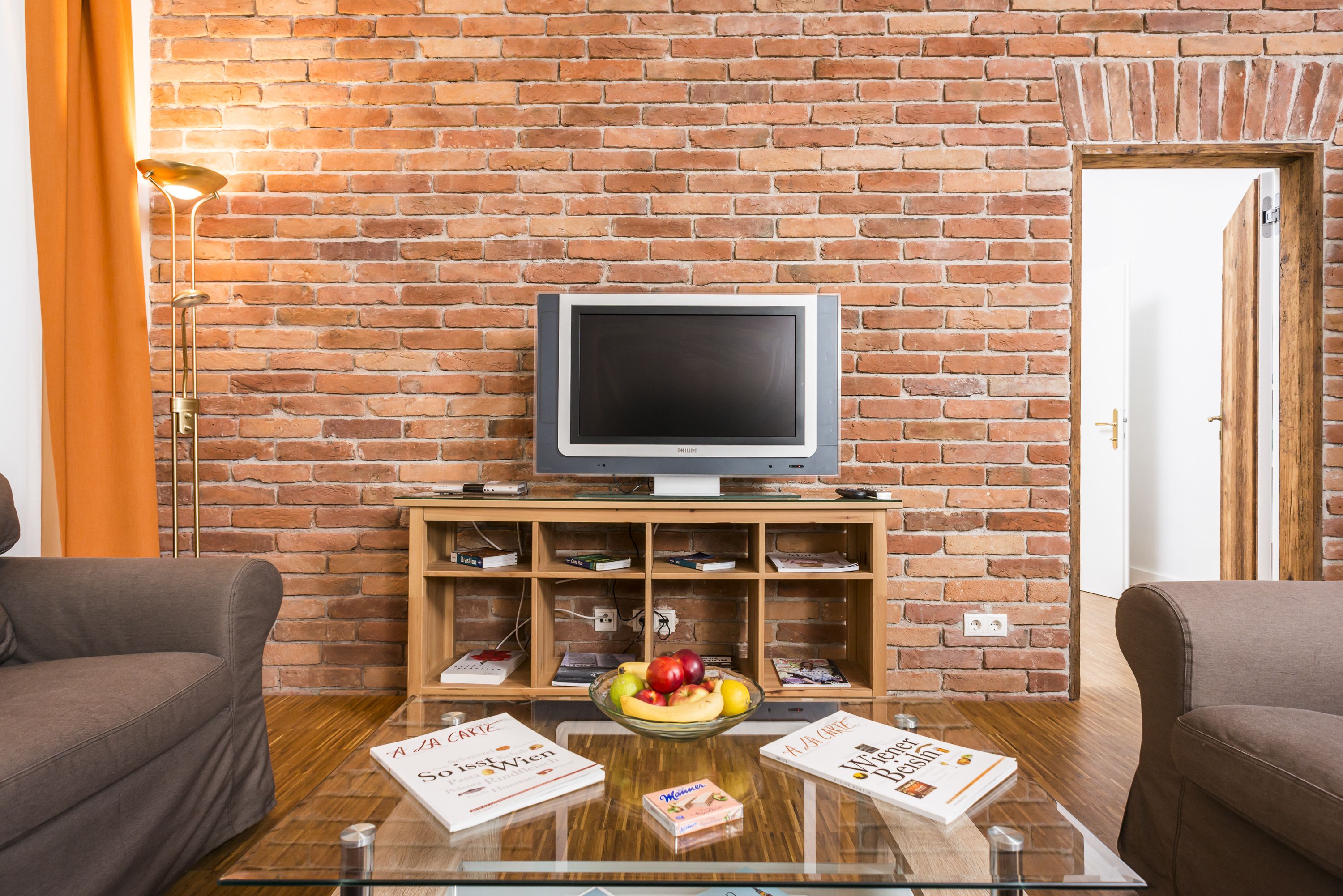 A living room with a brick wall, a flat-screen TV, a glass coffee table with books and fruit, and two sofas.