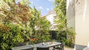An outdoor patio with a table and chairs, surrounded by greenery and a building in the background. - Appartements Ferchergasse (Vienna)