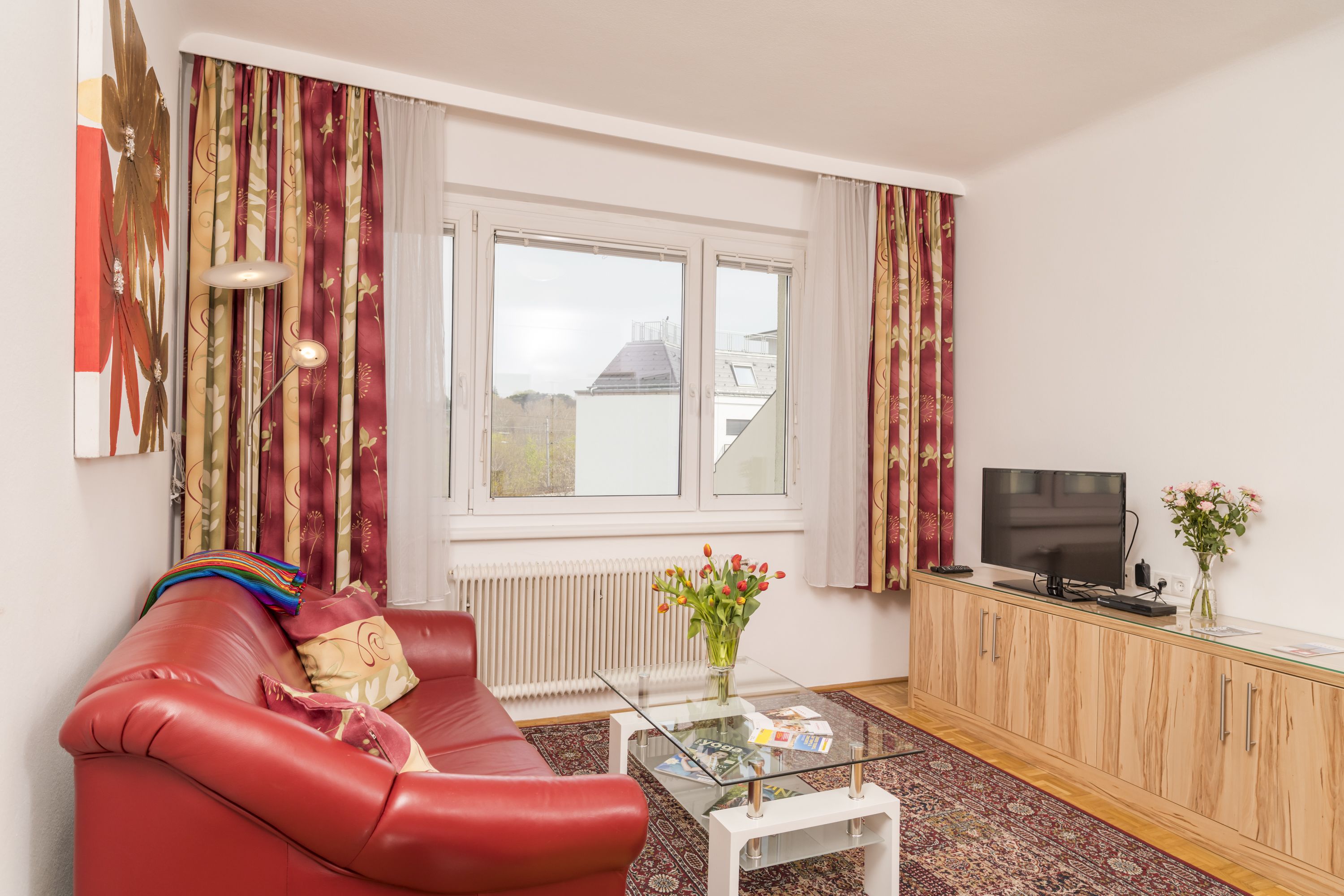 A living room with a red sofa, a glass coffee table, a TV on a wooden cabinet, and a window with curtains.