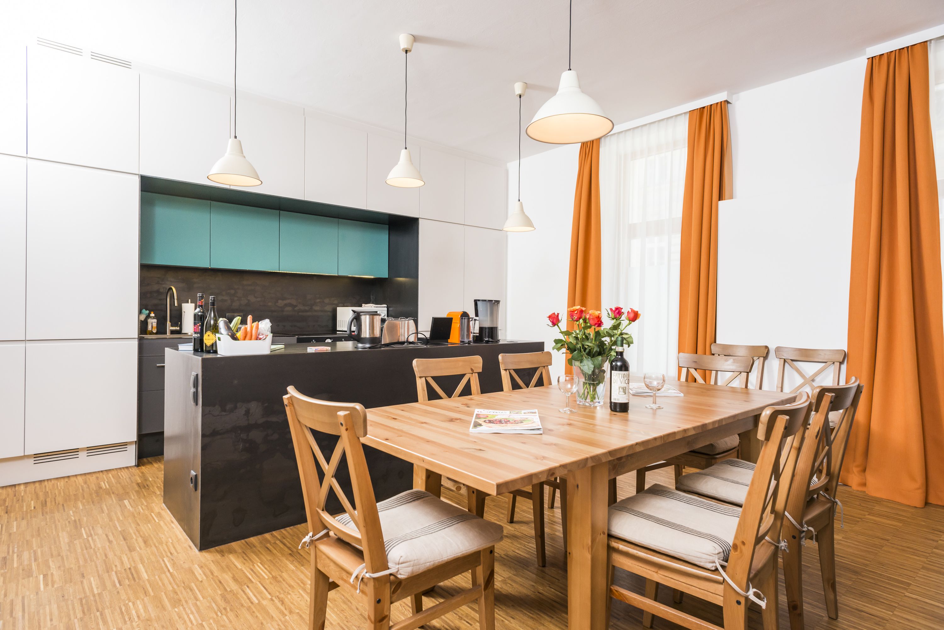 A modern kitchen with a central island, wooden dining table, and chairs. There are pendant lights above the island and a window with curtains.