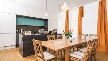 A modern kitchen with a central island, wooden dining table, and chairs. There are pendant lights above the island and a window with curtains.