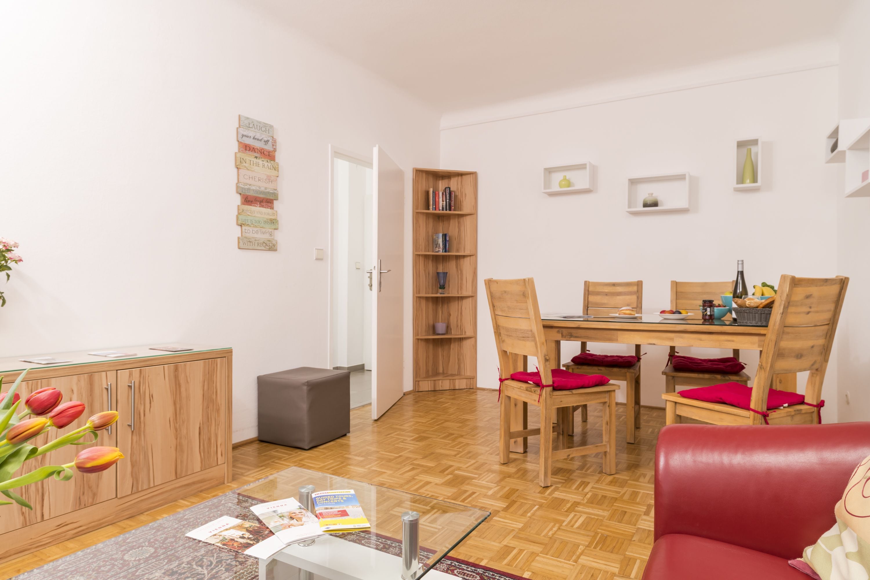 Een moderne woonkamer met een houten eettafel, rode fauteuils, een glazen salontafel en een boekenkast.