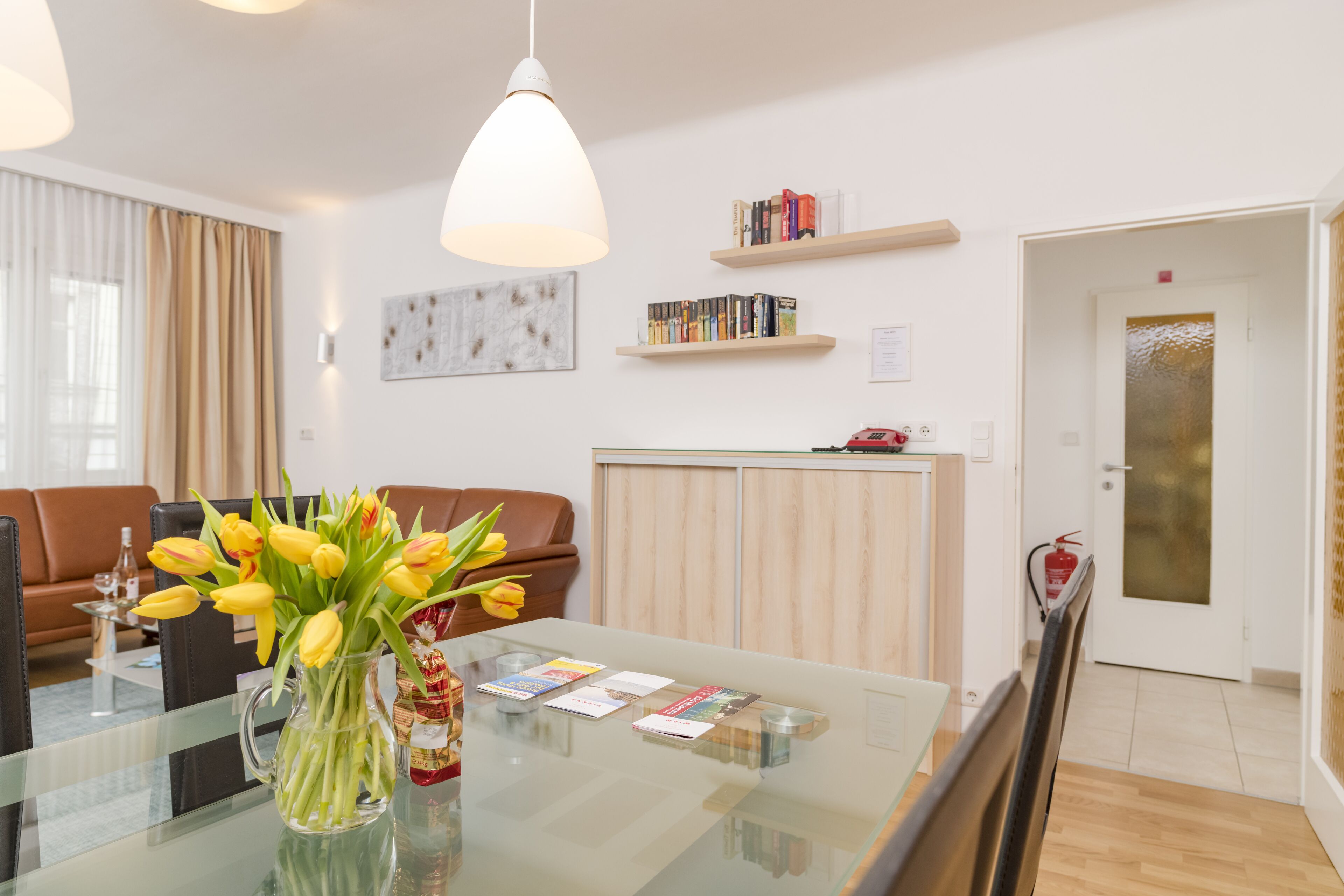 A modern living room with a glass coffee table, a vase of yellow tulips, a bookshelf, and a door with a frosted glass panel.