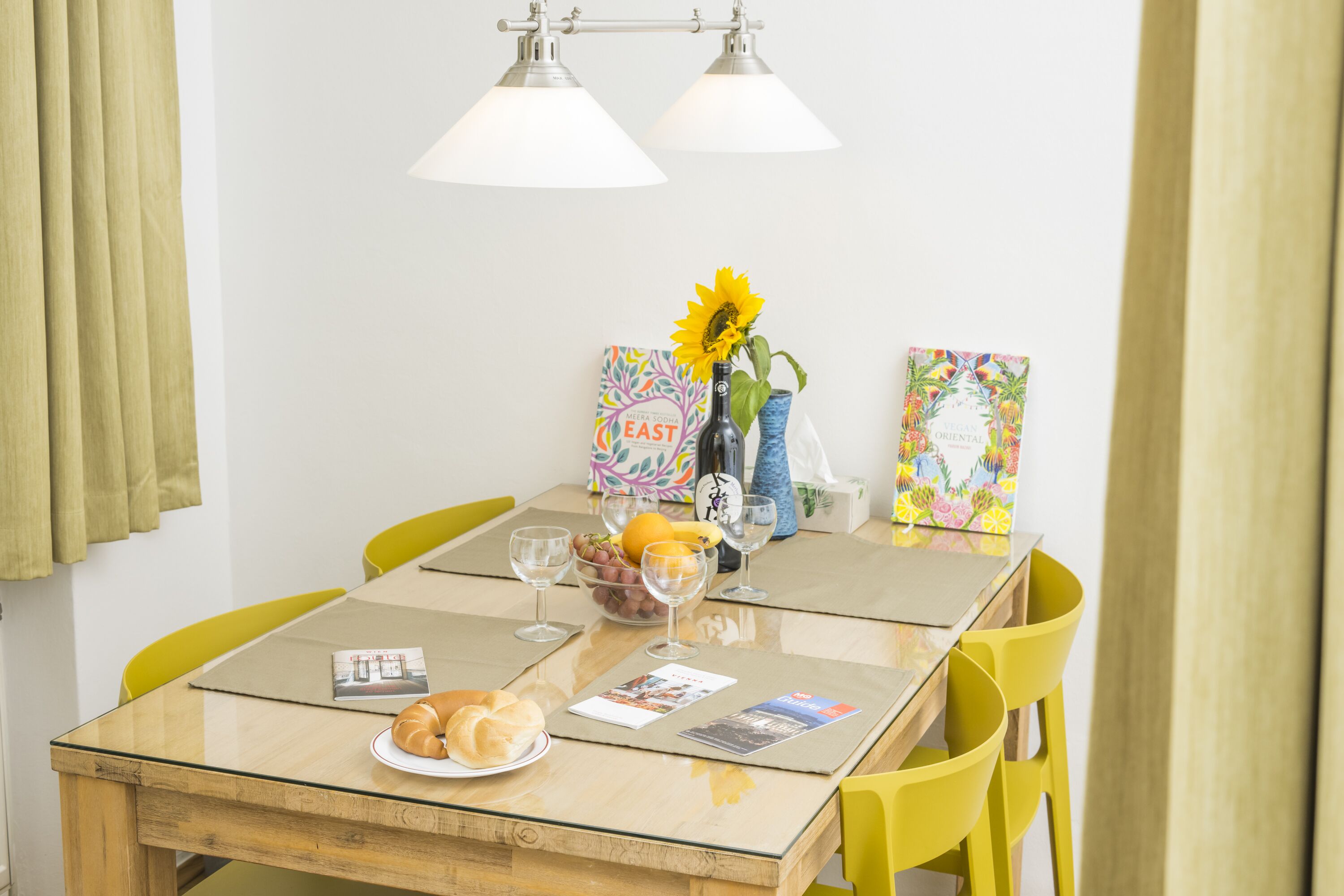 Een eettafel met een glazen blad, omringd door gele stoelen, met een fruitschaal, een fles en een vaas met een zonnebloem.