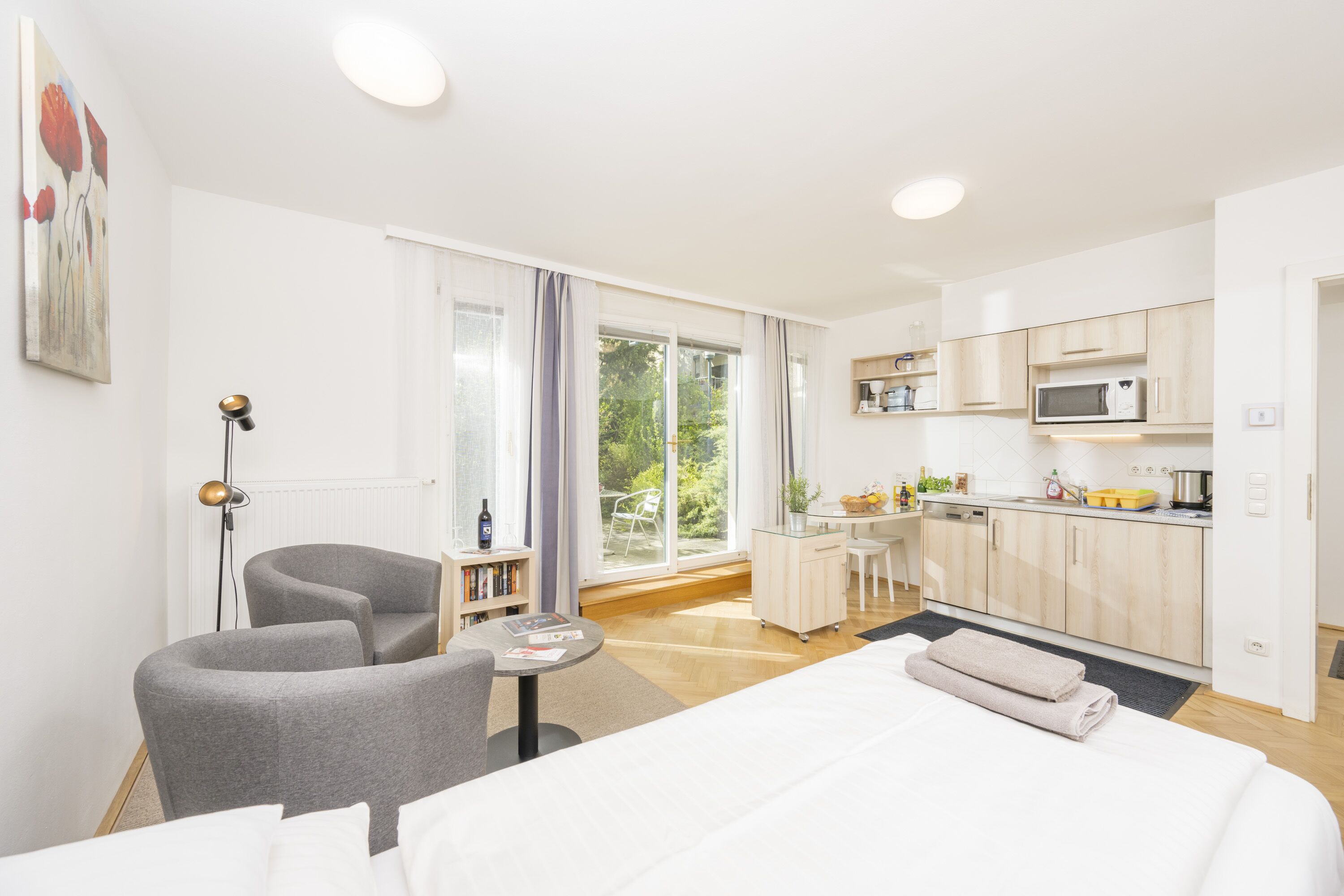 A modern hotel room with a bed, a desk, a chair, a sofa, and a kitchenette.
