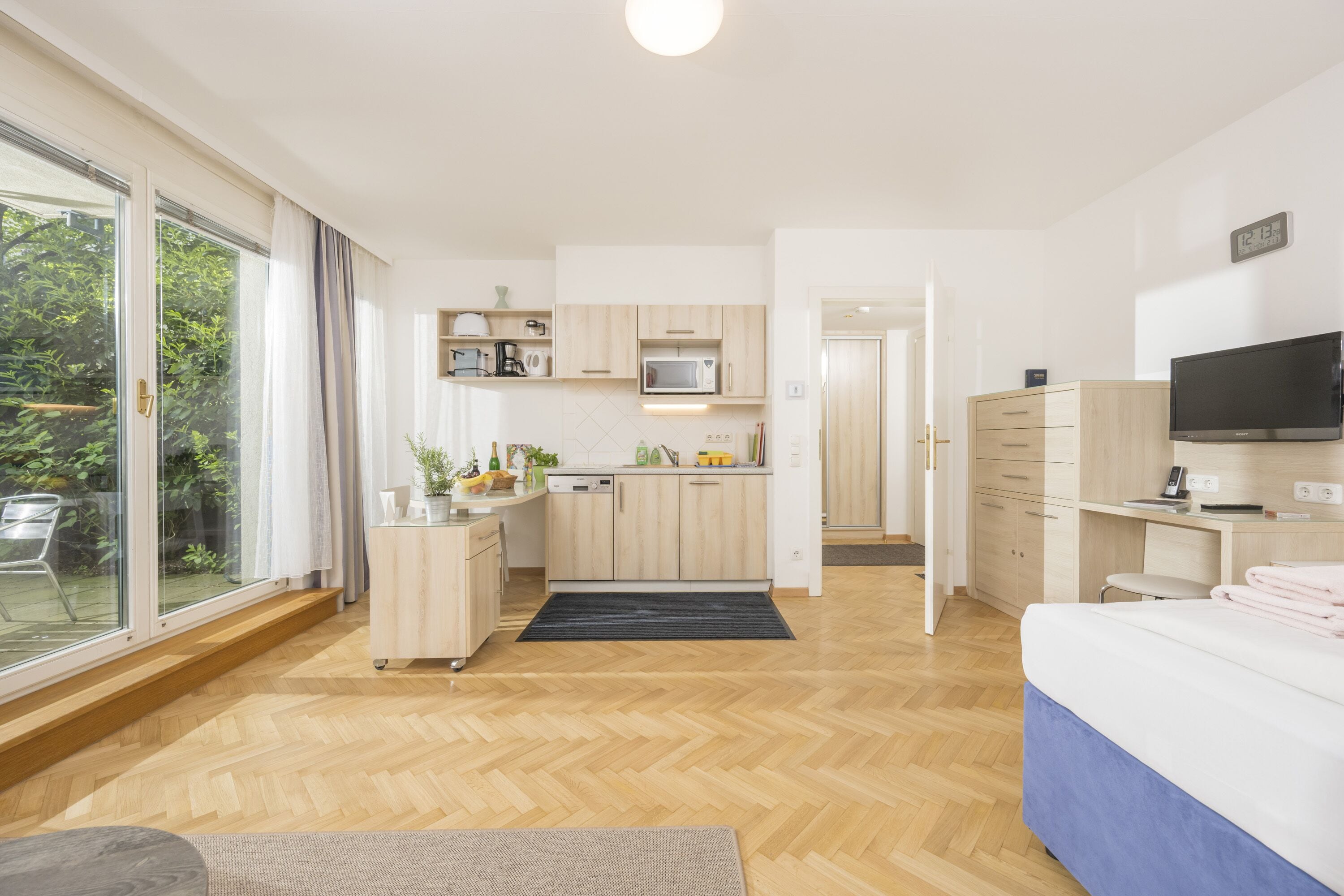Moderni hotellihuone, jossa on suuri sänky, televisio, työpöytä ja pieni keittiö.