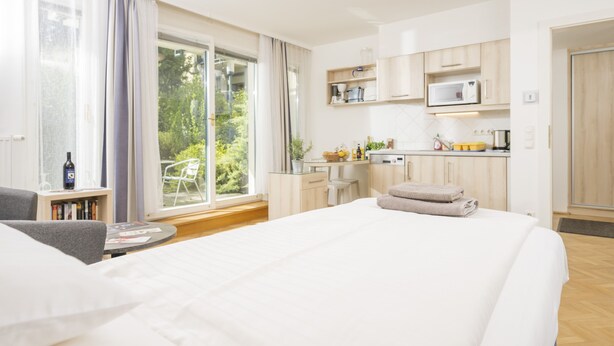 A hotel room with a large bed, a dining table, a kitchen area, and a view of the outdoors. - Appartements Ferchergasse (Vienna)