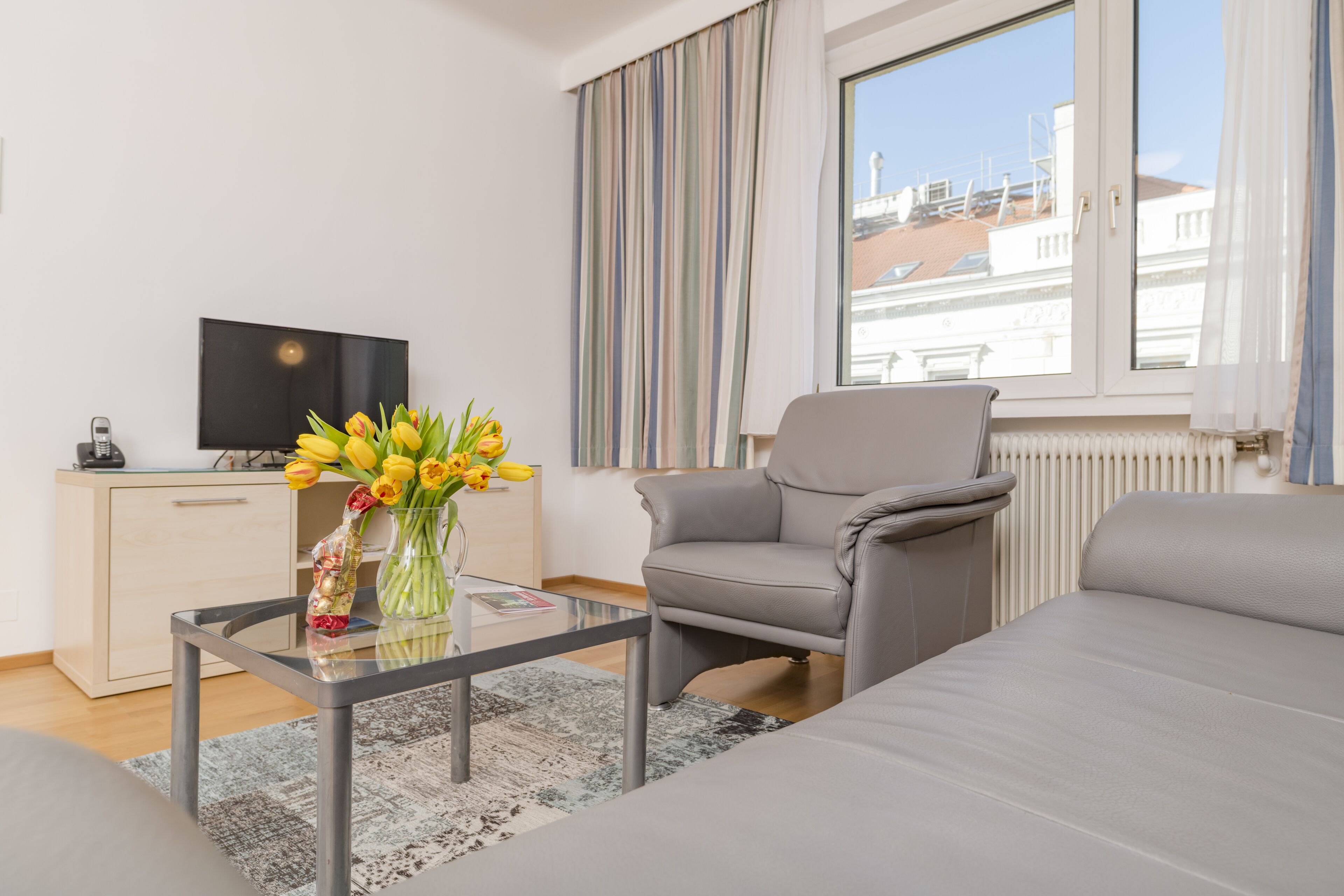 Ett modernt vardagsrum med en grå soffa, ett soffbord i glas med en vas med blommor, en tv-apparat placerad på ett träskåp och en utsikt över byggnader genom ett fönster med randiga gardiner.