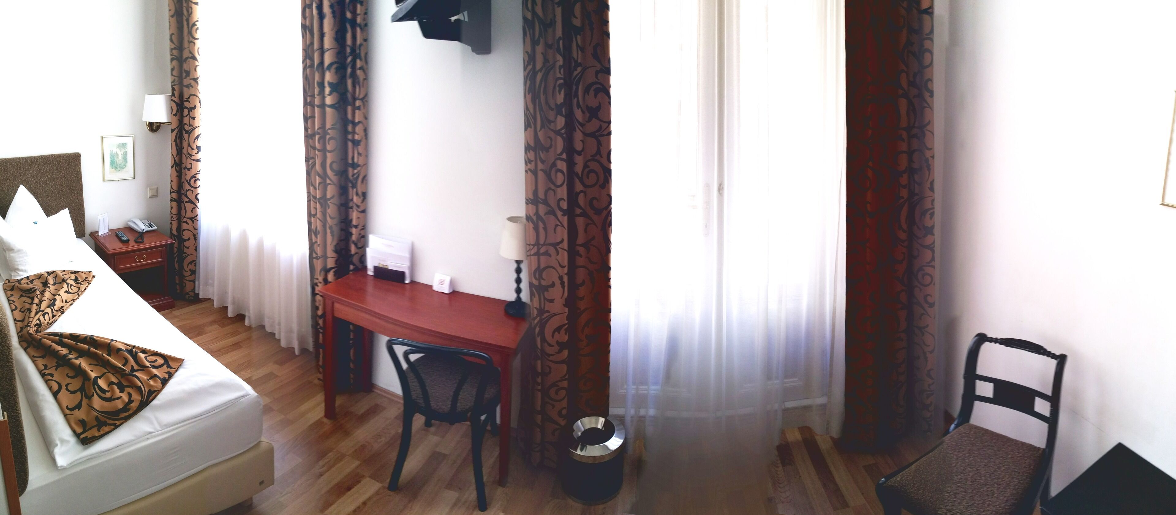 A hotel room with a bed, a desk, a chair, a waste bin, and curtains.