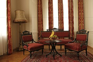 Una habitación con dos sillones tapizados de color rojo, una mesita redonda con fruteros, una lámpara de pie y una alfombra estampada.