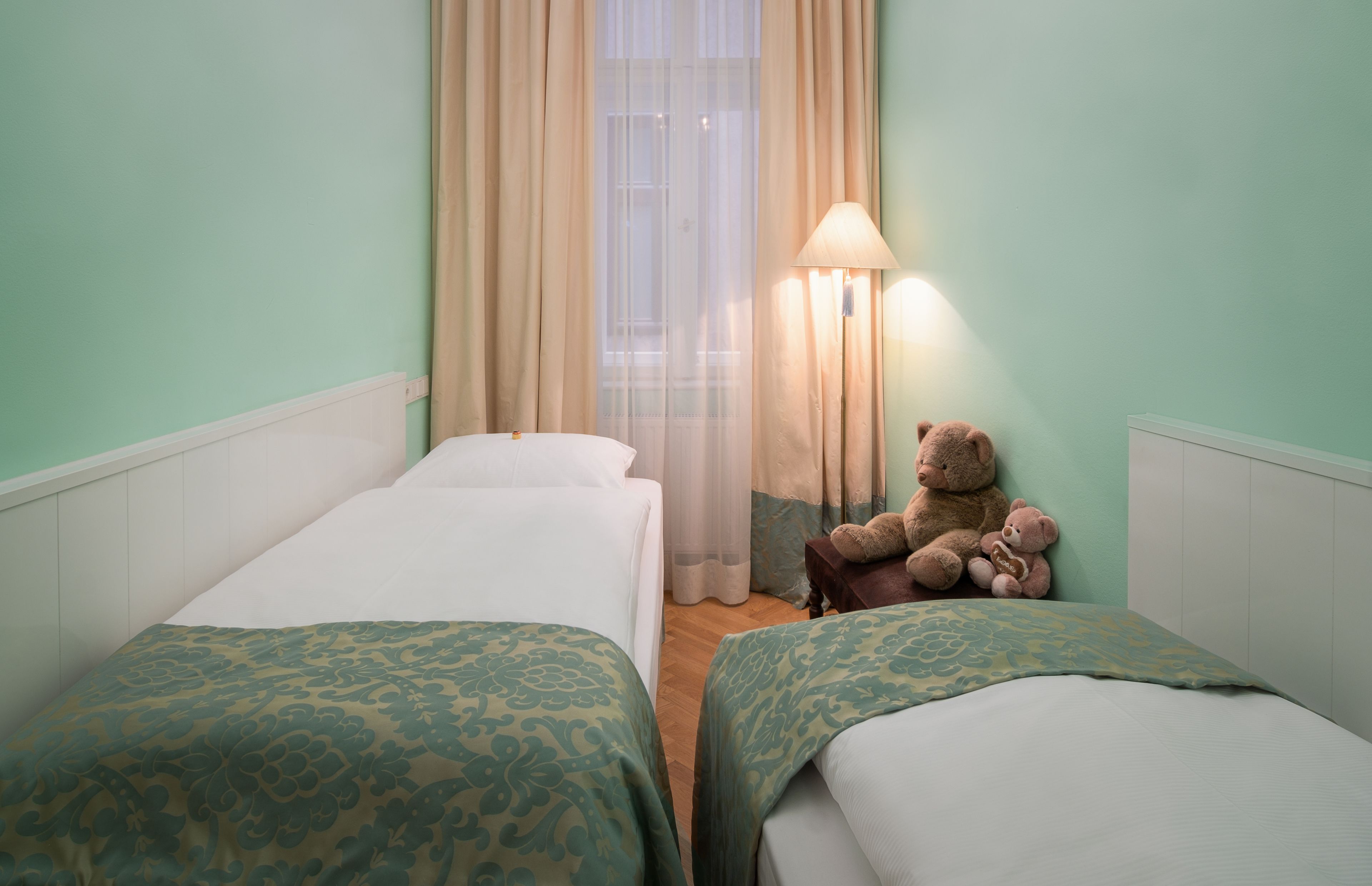A hotel room with two single beds, a lamp, a teddy bear, and a window with curtains.