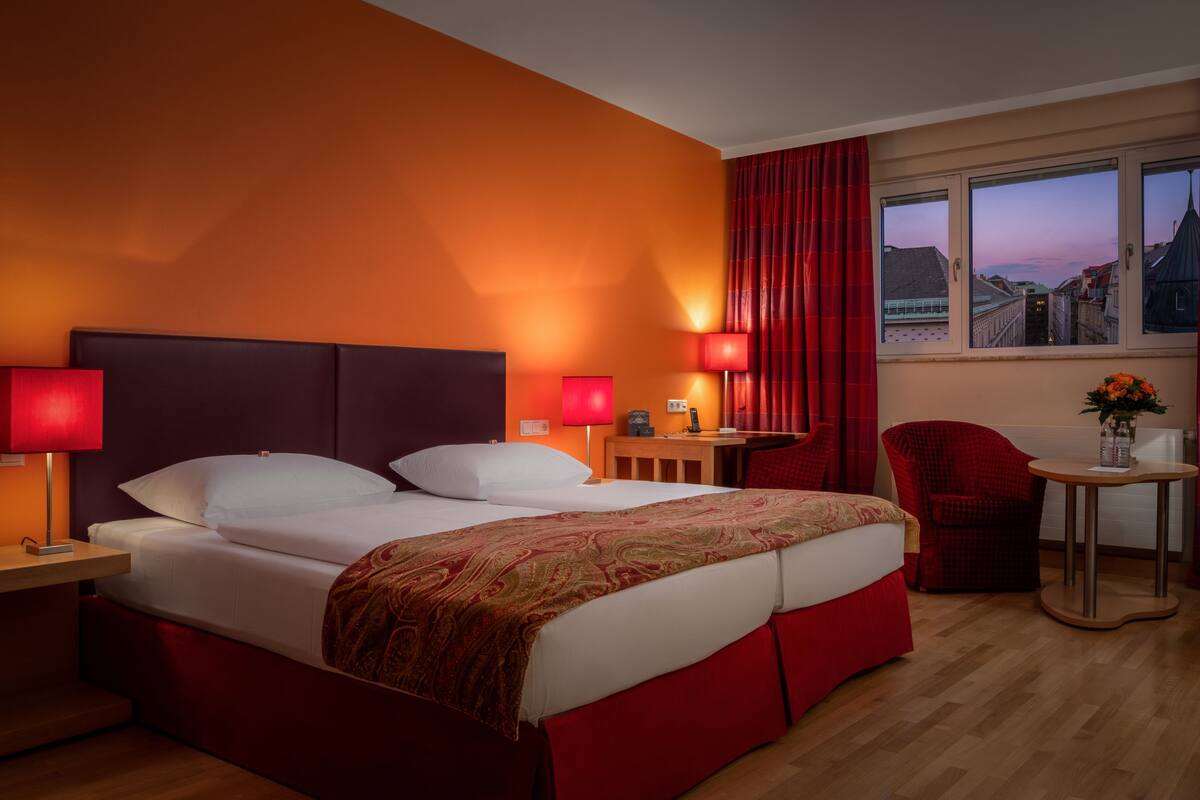 A hotel room with a large bed, two bedside tables with lamps, a small table with a chair, and a window with a city view.
