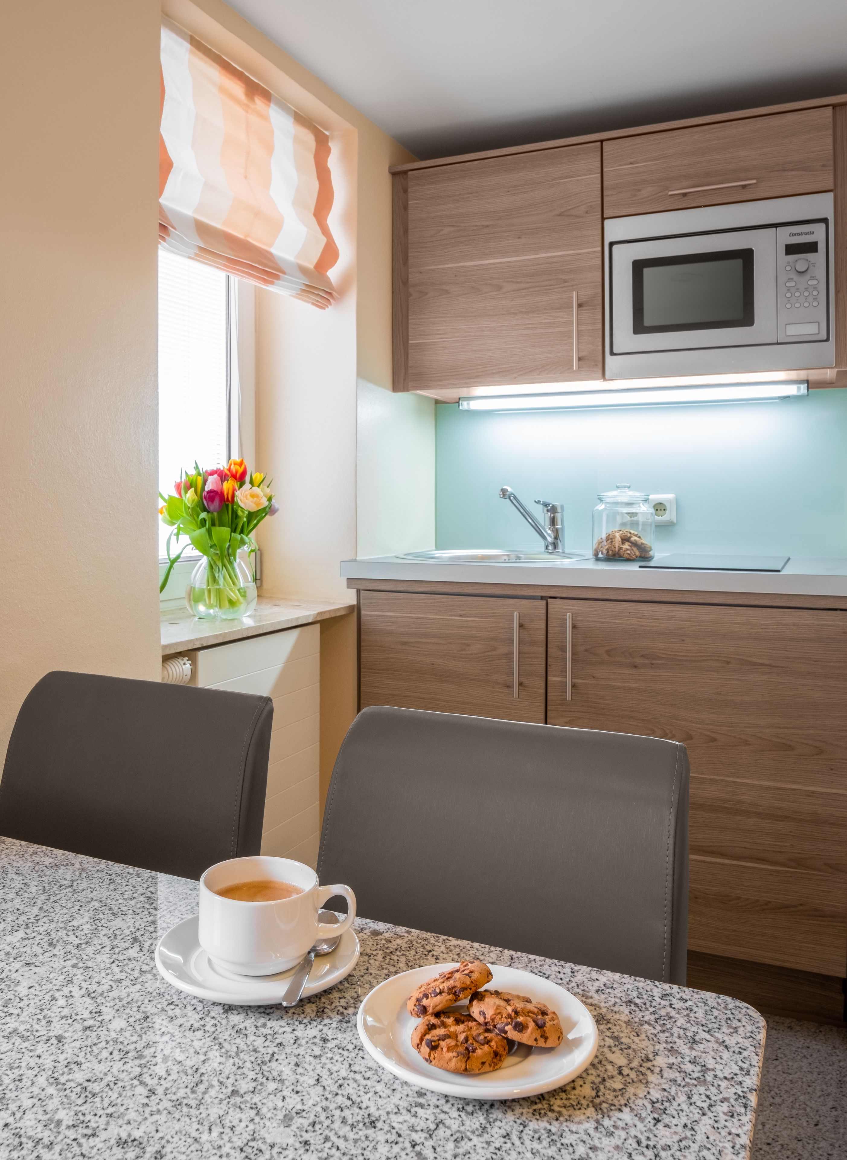 Uma cozinha compacta com micro-ondas, pia e armários de madeira. Há uma área de refeições com mesa, cadeiras e um prato de biscoitos. Uma xícara de café está sobre a mesa, e um vaso com flores repousa no parapeito da janela.