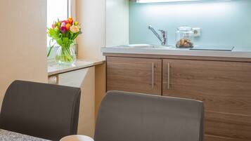 Uma cozinha compacta com micro-ondas, pia e armários de madeira. Há uma área de refeições com mesa, cadeiras e um prato de biscoitos. Uma xícara de café está sobre a mesa, e um vaso com flores repousa no parapeito da janela.