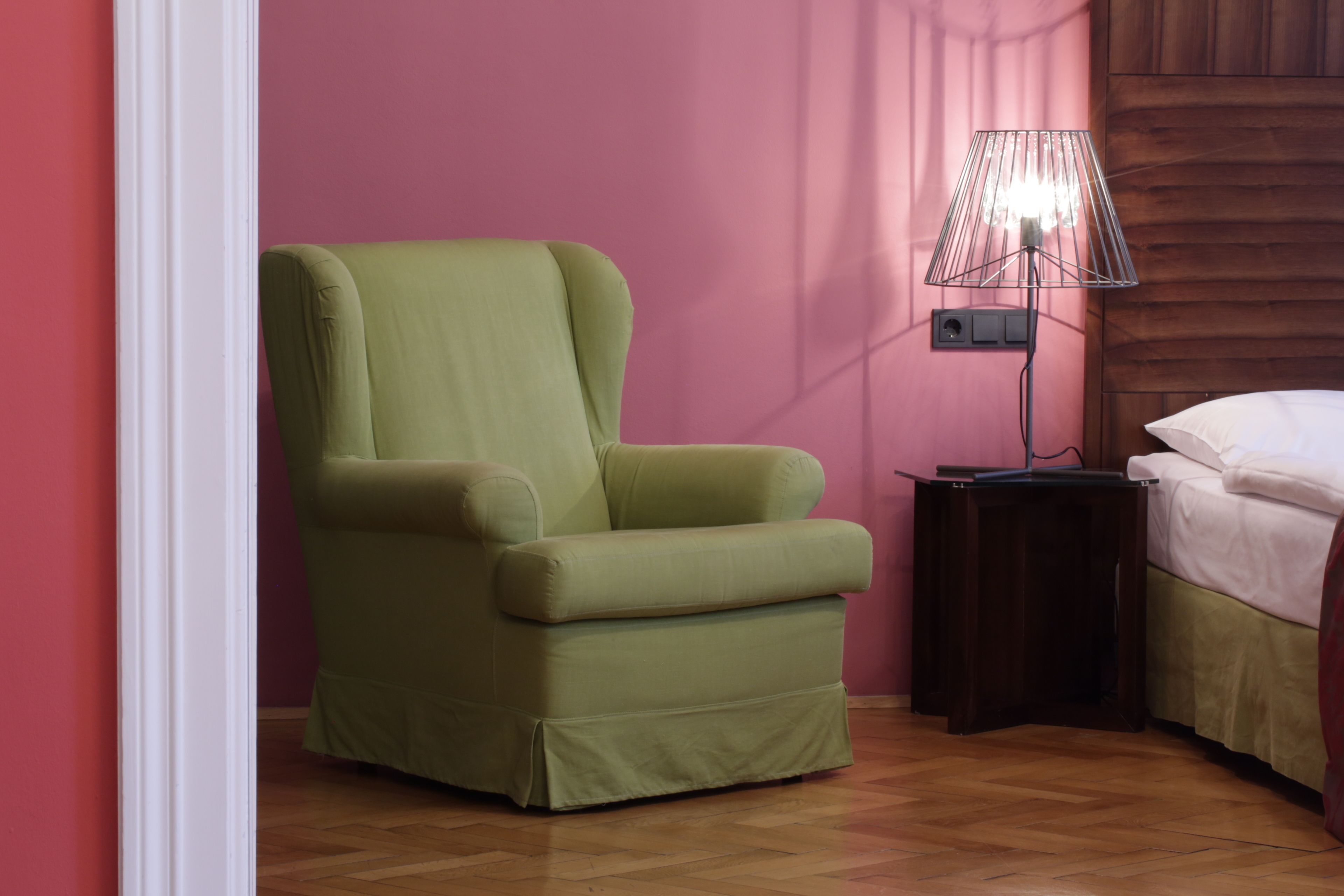 Una habitación con un sillón verde, una mesita de noche con lámpara y una cama con ropa de cama blanca.