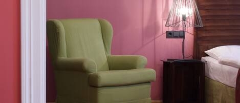 Una habitación con un sillón verde, una mesita de noche con lámpara y una cama con ropa de cama blanca.