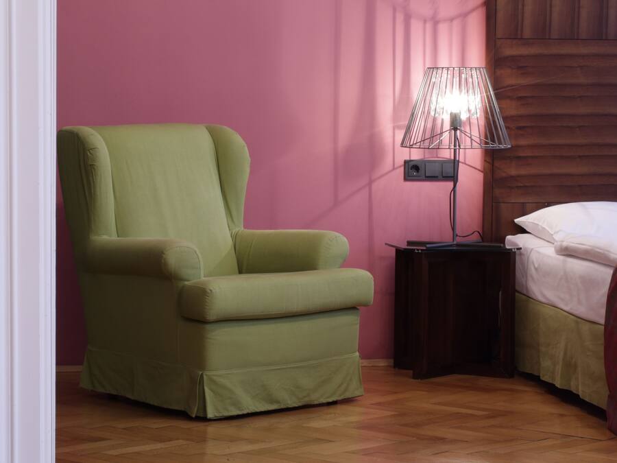 Una habitación con un sillón verde, una mesita de noche con lámpara y una cama con ropa de cama blanca.