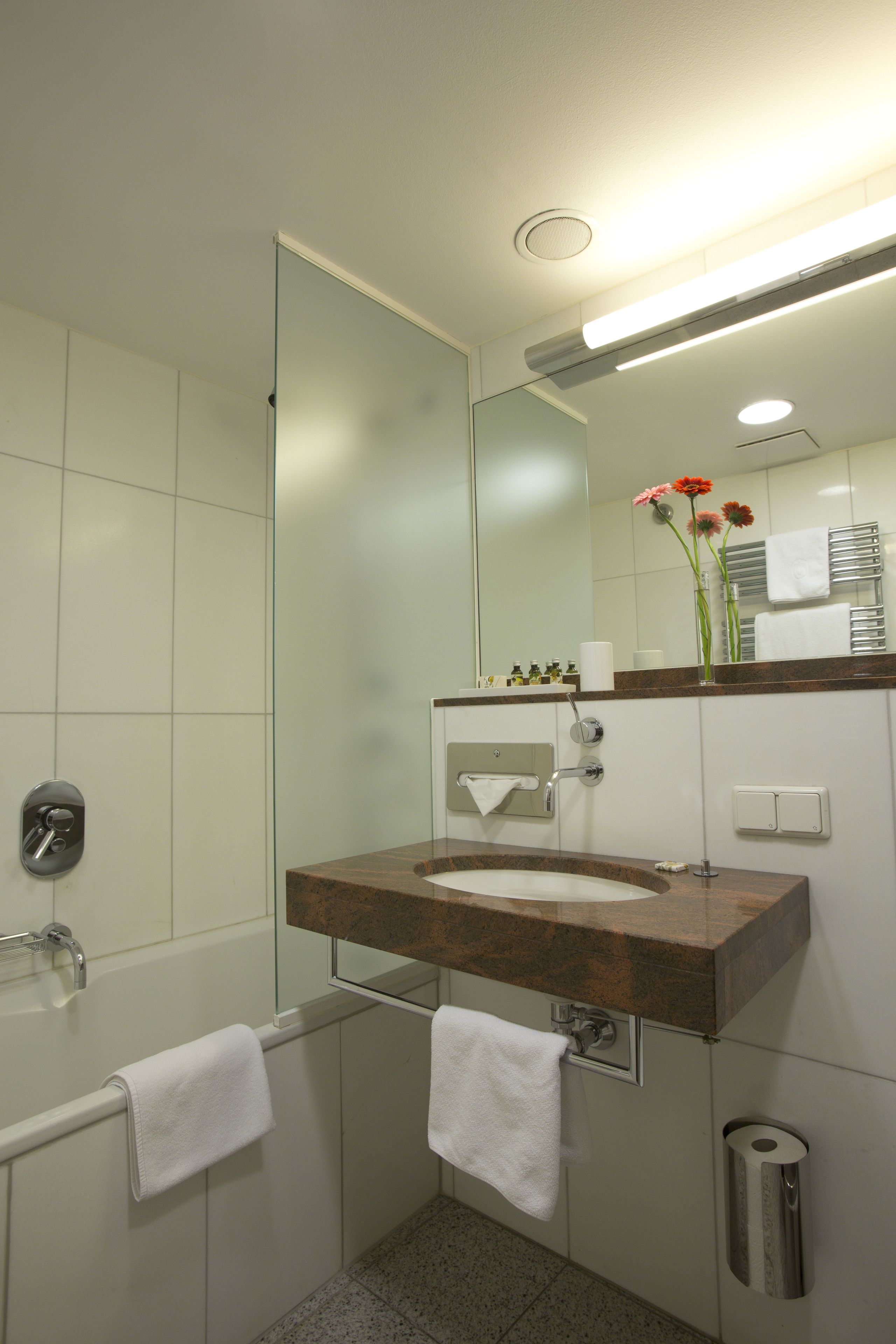 Un baño moderno con lavamanos de mármol, cabina de ducha de vidrio y un espejo que refleja un jarrón con flores.