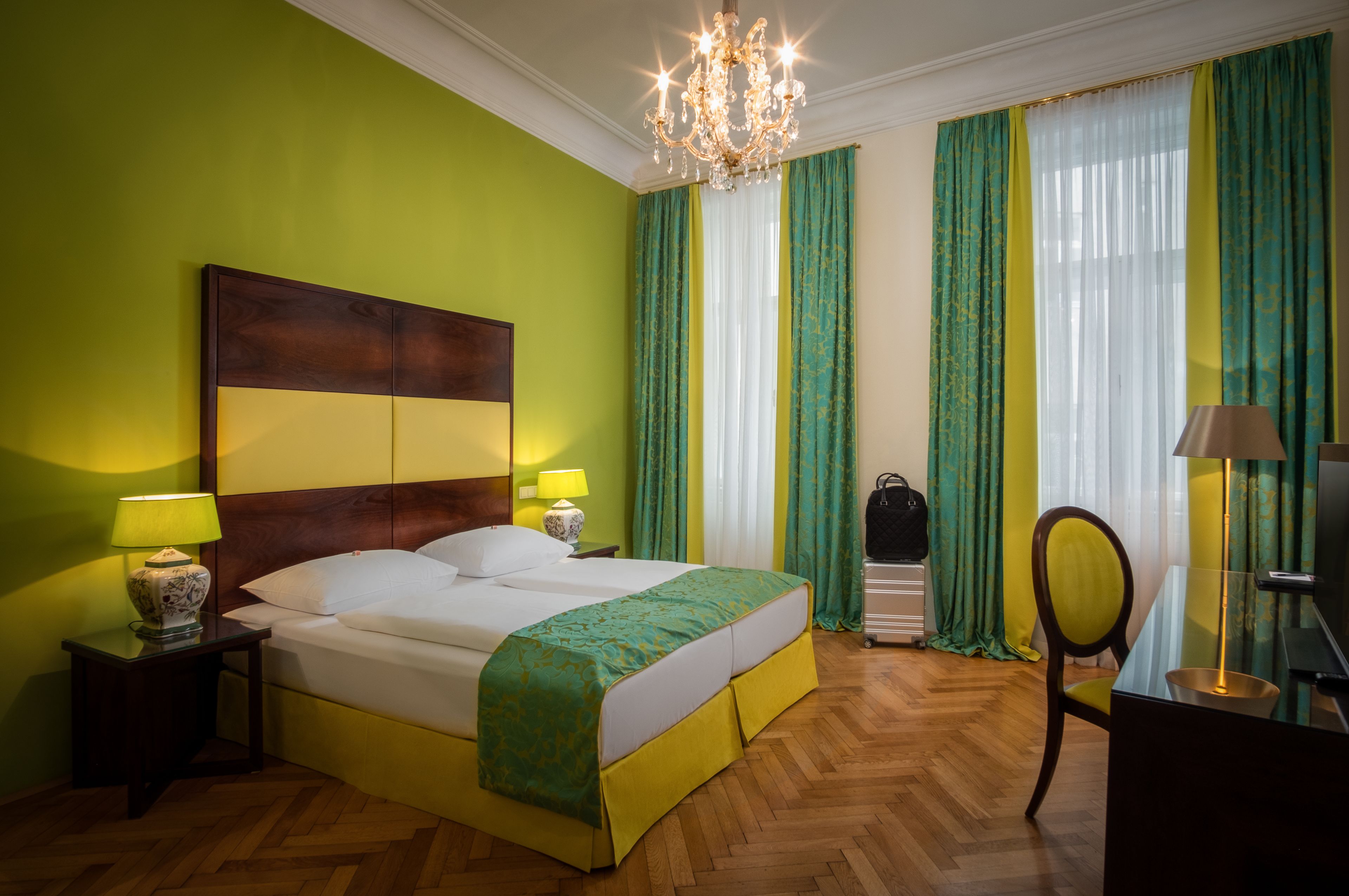 A hotel room with a large bed, two bedside lamps, a chandelier, a desk with a lamp, and a chair.