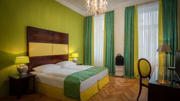 A hotel room with a large bed, two bedside lamps, a chandelier, a desk with a lamp, and a chair.