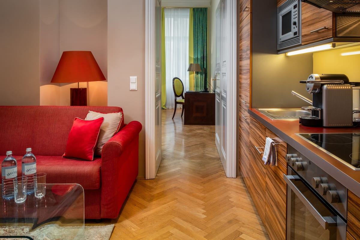 A modern hotel room with a red sofa, a glass coffee table, a lamp, and a kitchenette with a microwave and stove.