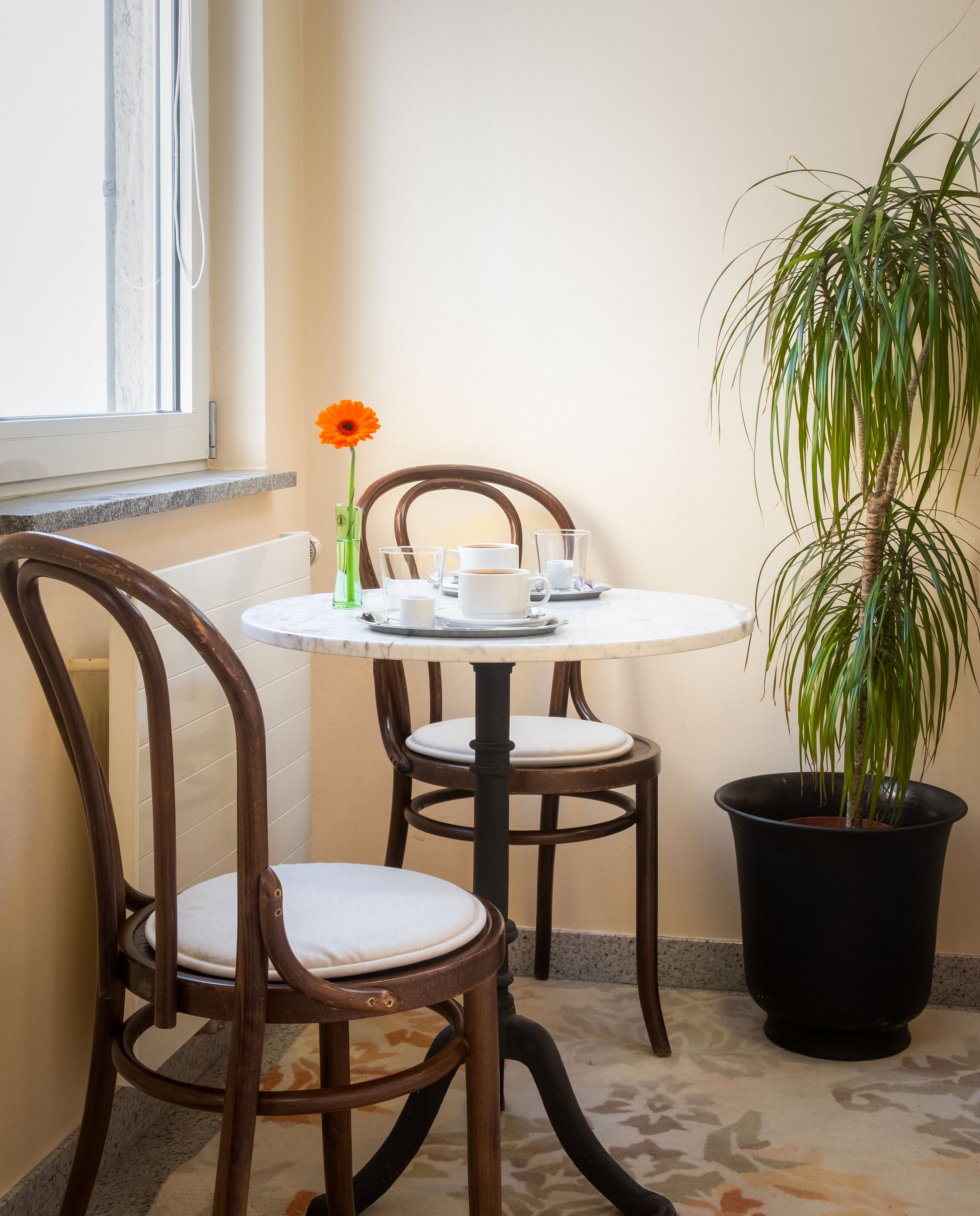 A small round table with a white tablecloth, two chairs, a potted plant, and a vase with a single orange flower.