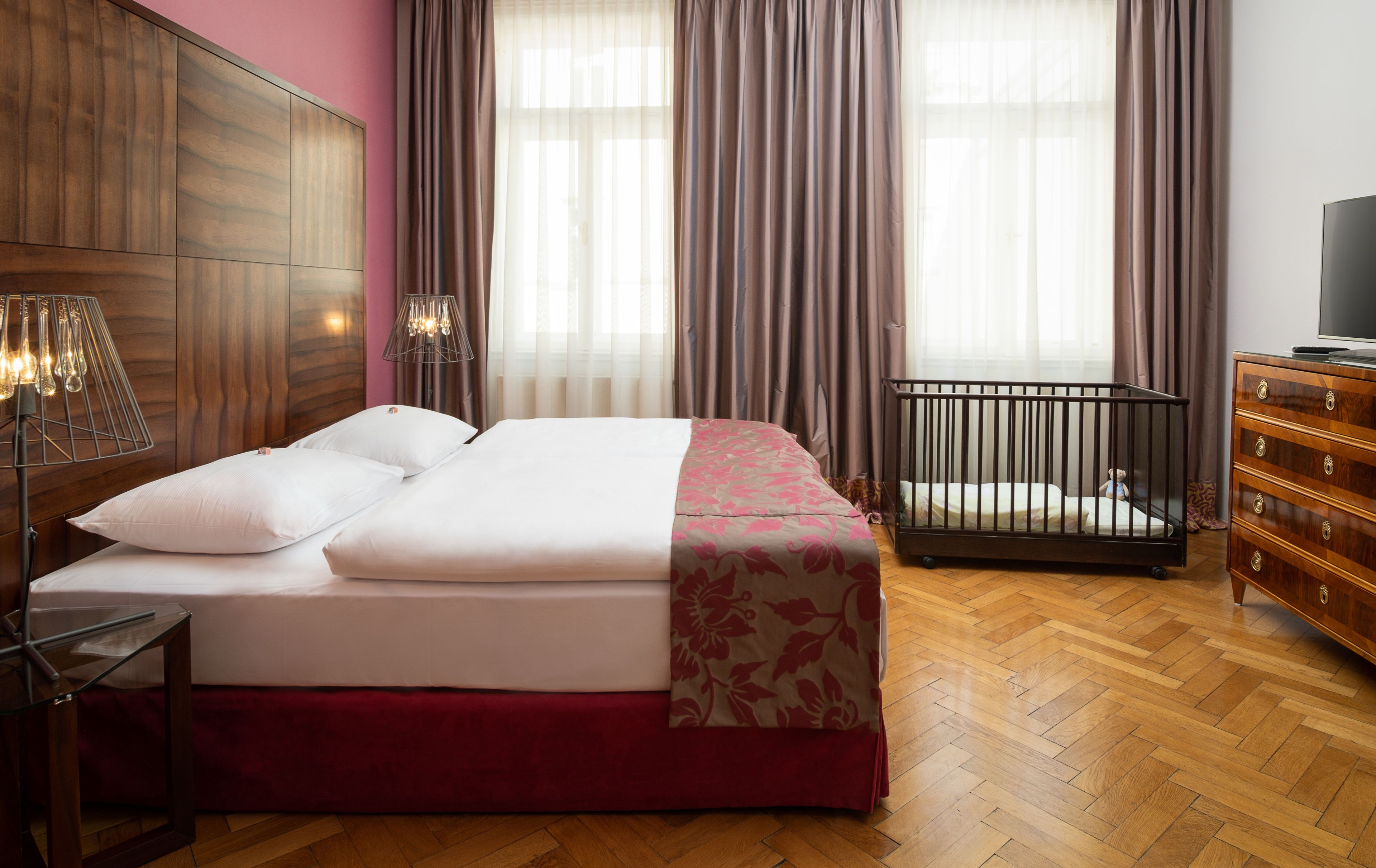 A hotel room with a wooden headboard, a bed with white and pink bedding, a crib, a wooden dresser, and a television.