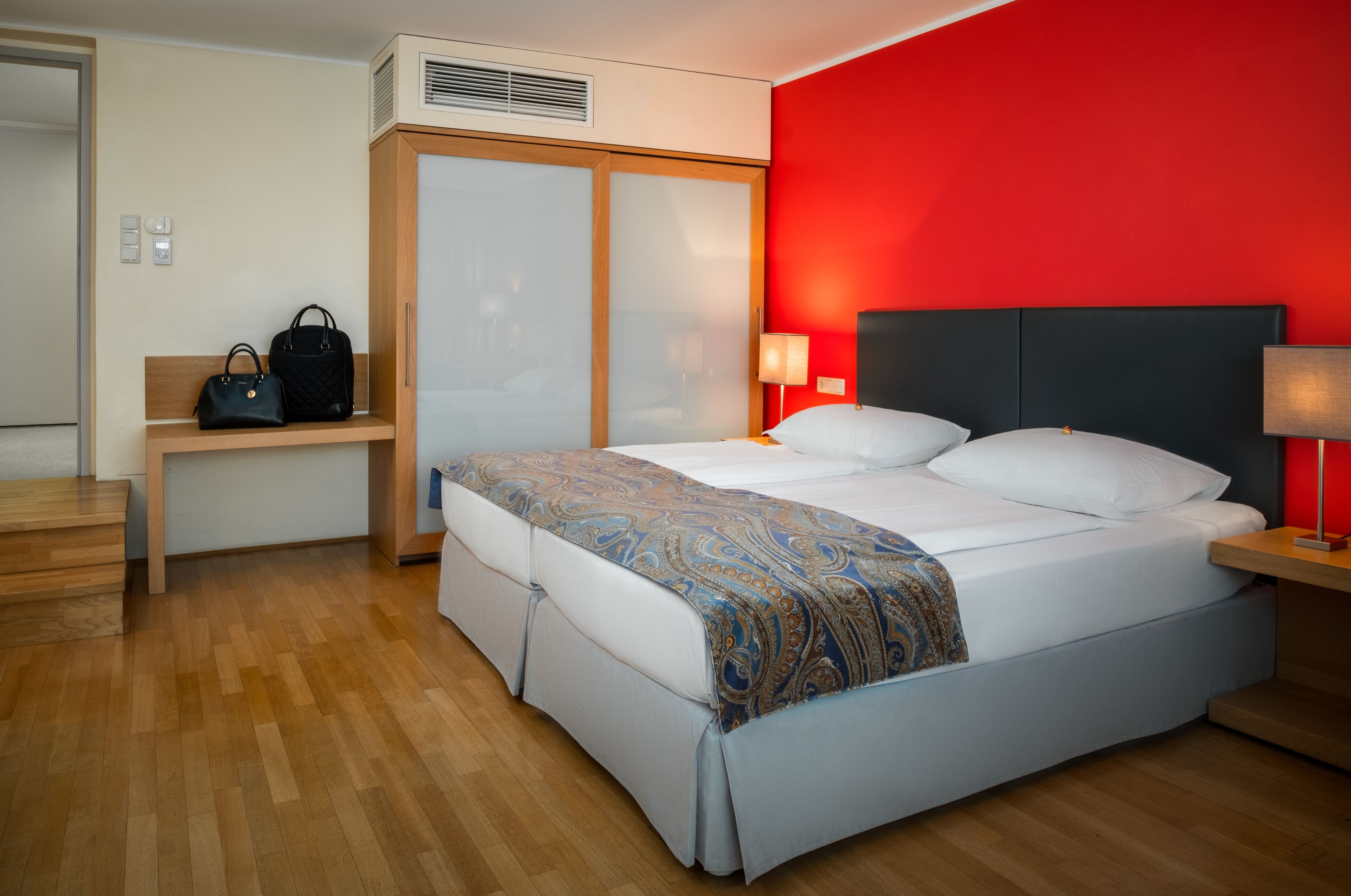 A hotel room with a bed, bedside tables, a nightstand, a wardrobe, and a bench with handbags.